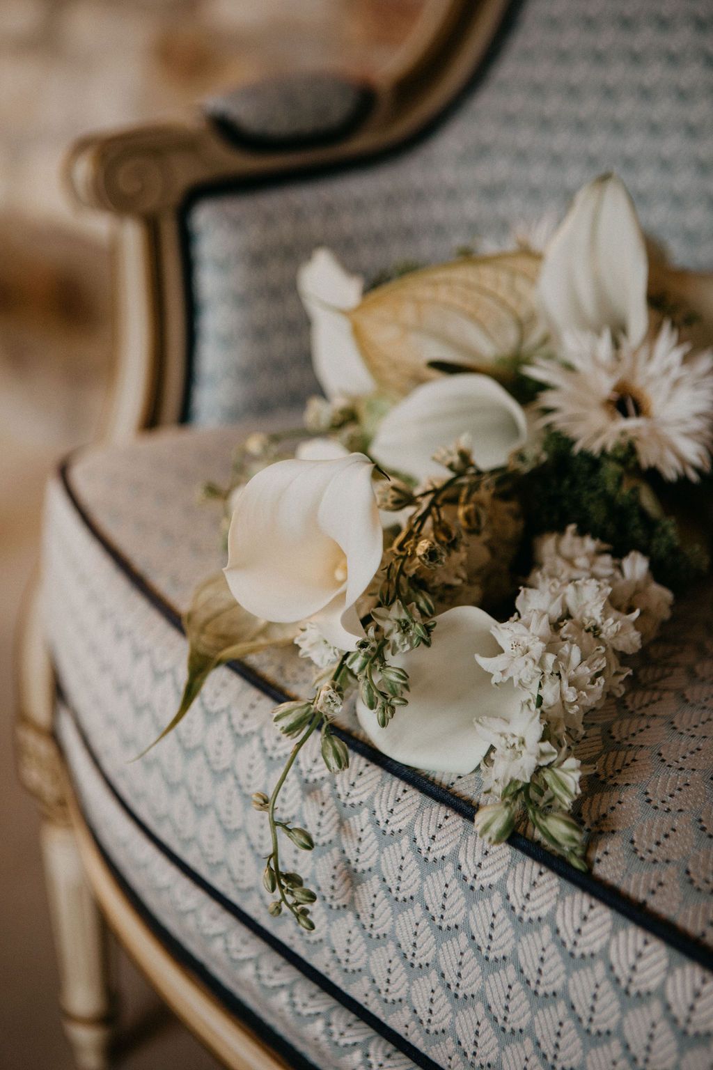 Elegant white wedding bouquet with calla lilies resting on a vintage French upholstered chair.