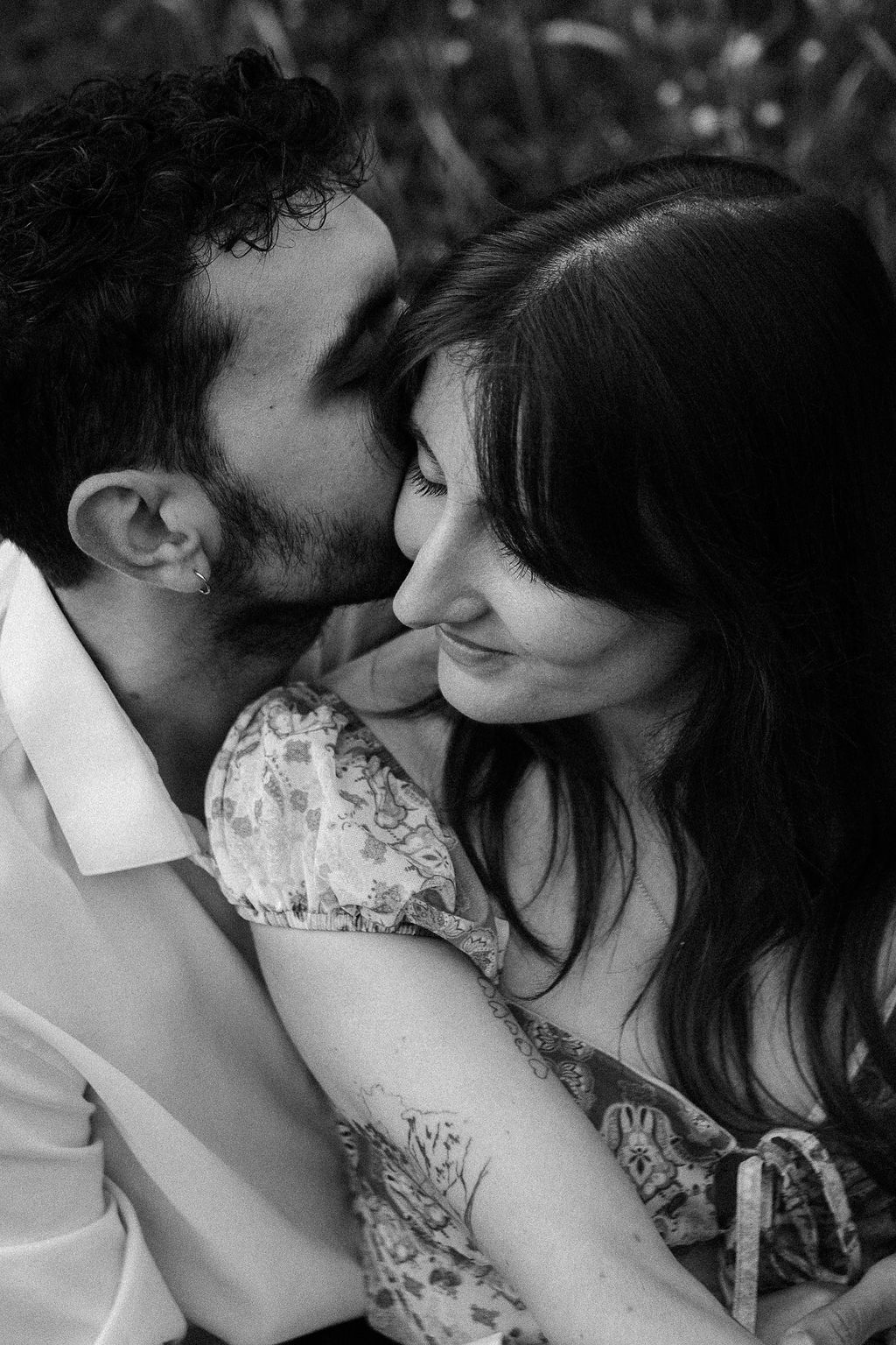 Black and white portrait of man kissing woman on cheek as she smiles in a garden setting.