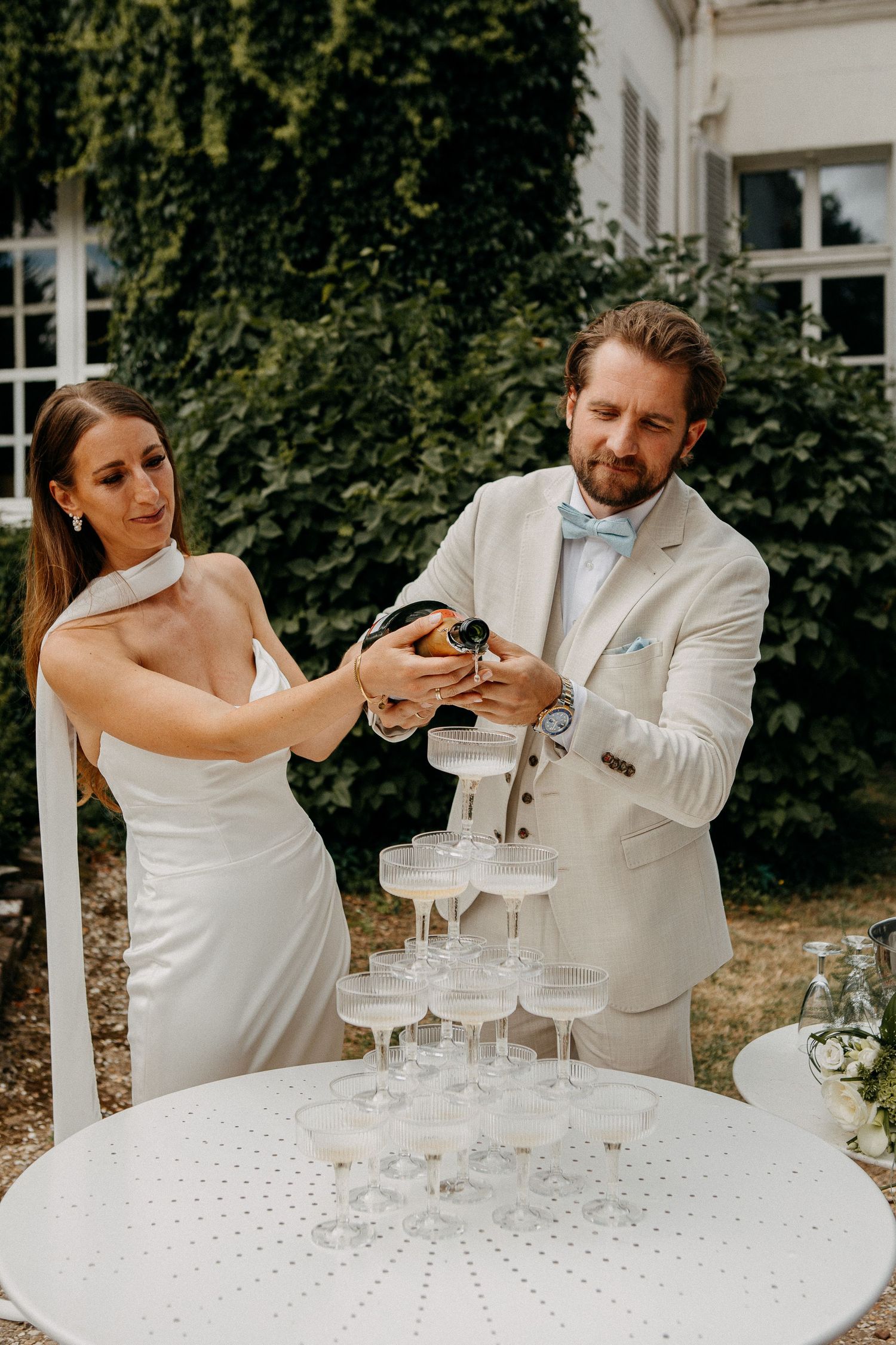 Bride and groom pouring champagne tower at outdoor wedding reception in white attire.