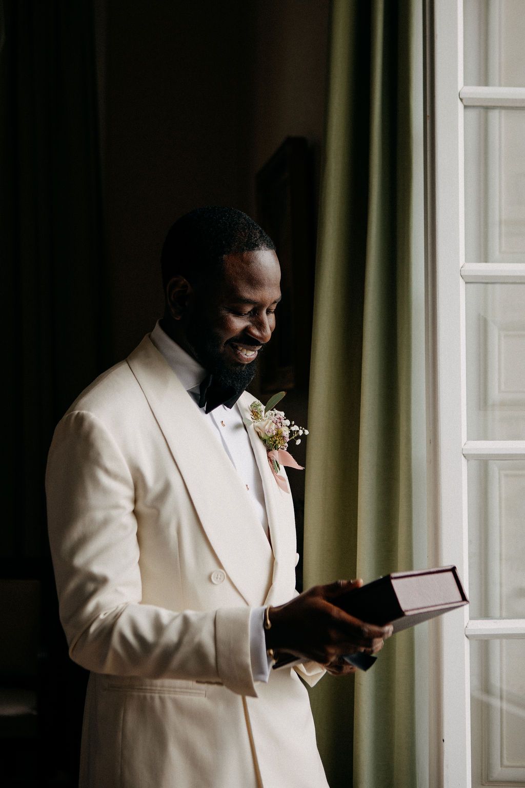 A joyful groom in a cream tuxedo reads his wedding vows by a sunlit window.