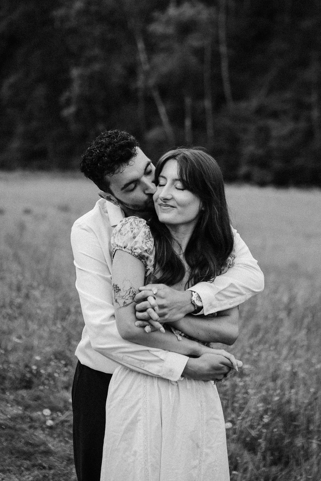Black and white photo of a man embracing and kissing a smiling woman in a meadow setting.
