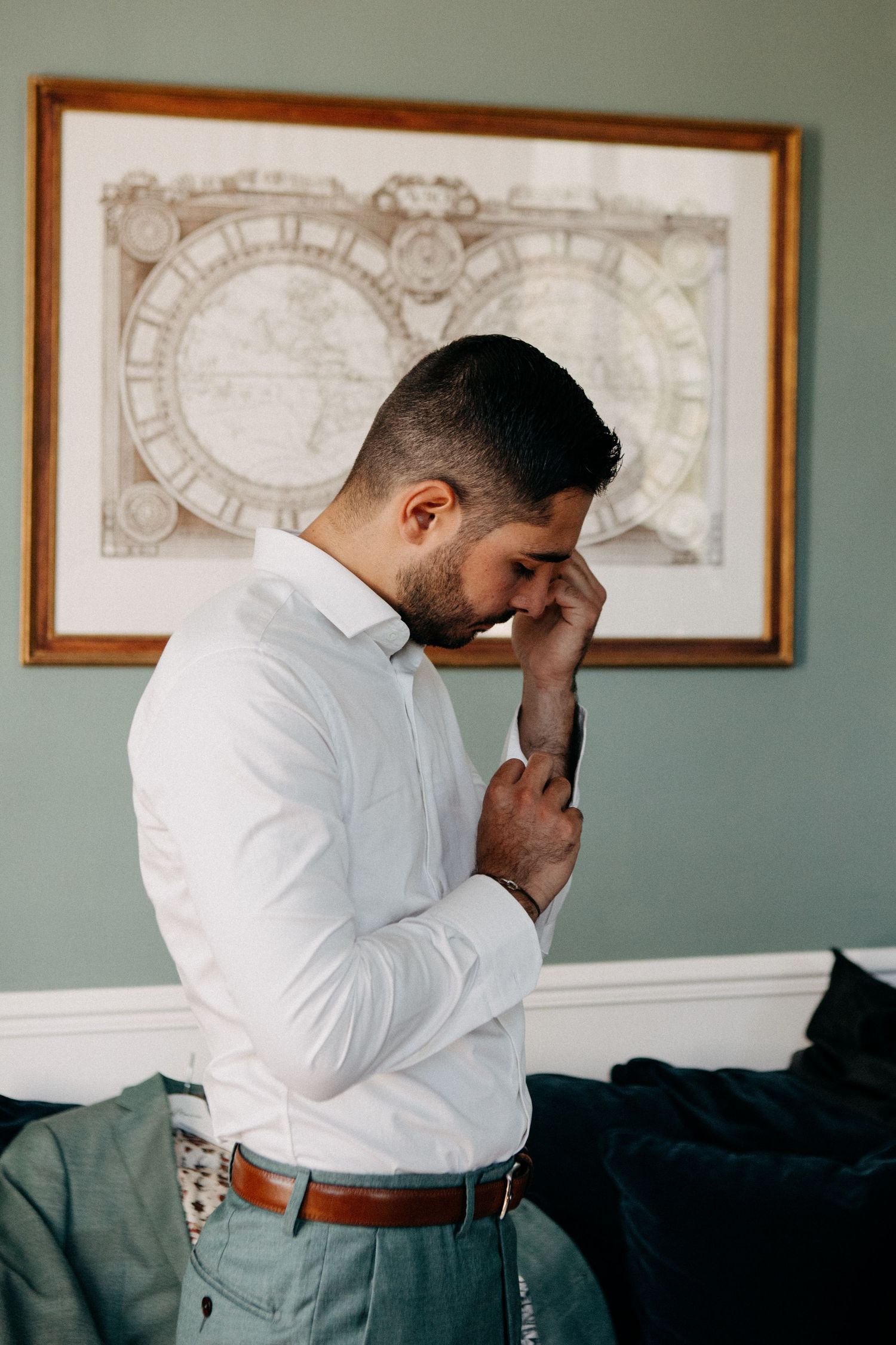 Groom in white dress shirt buttoning cuffs while getting ready in room with vintage map wall art.
