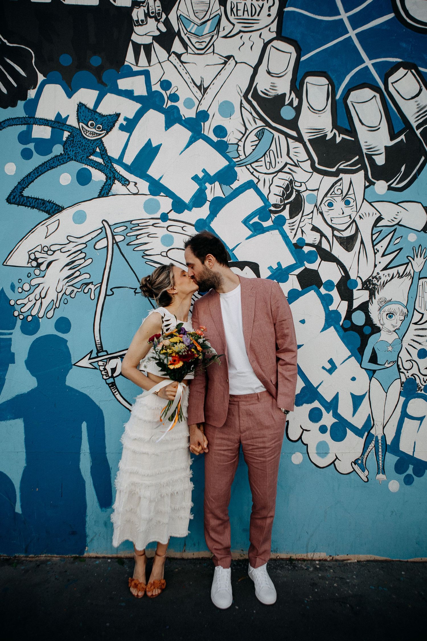 Bride and groom kissing in front of a vibrant blue graffiti mural, she in white fringe dress, he in pink suit.