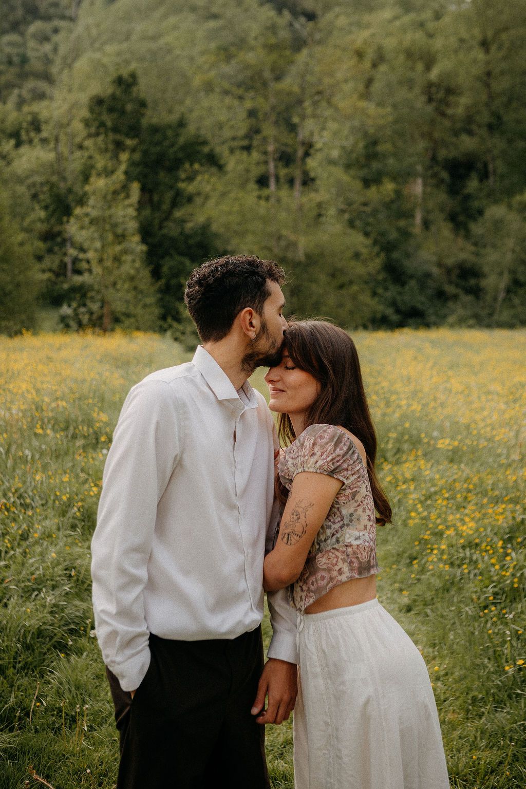 Man kissing woman on forehead in a yellow wildflower field with green forest behind them.