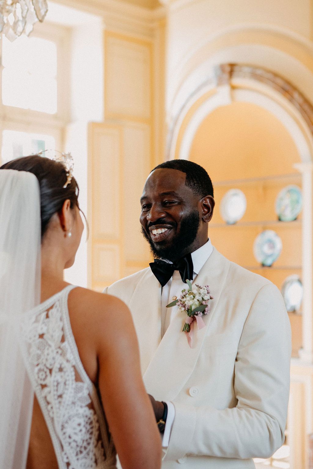 A beaming groom in a cream tuxedo smiles warmly at his bride during their wedding ceremony.