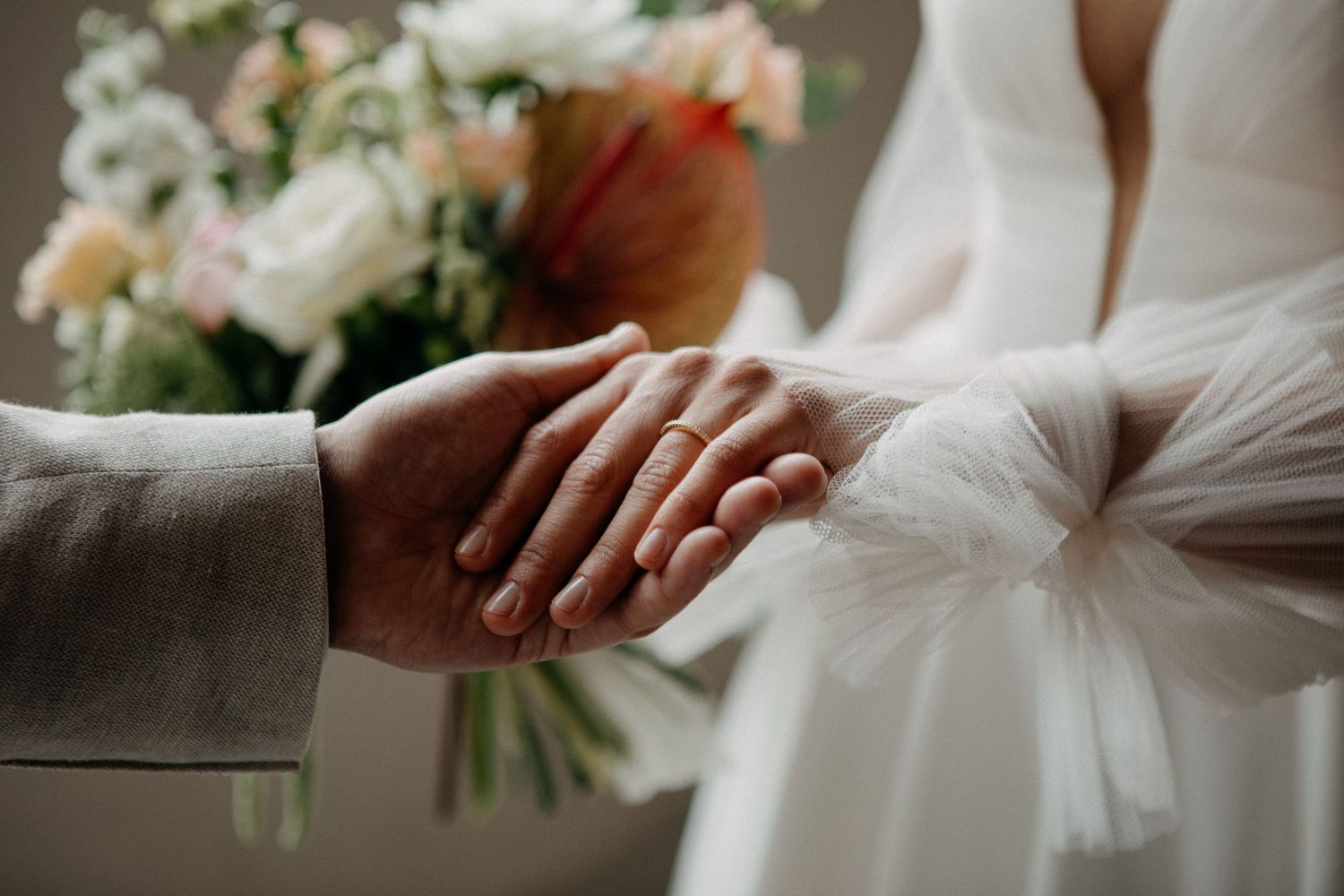 Close-up of bride and groom holding hands, with a colorful bridal bouquet softly blurred in background.