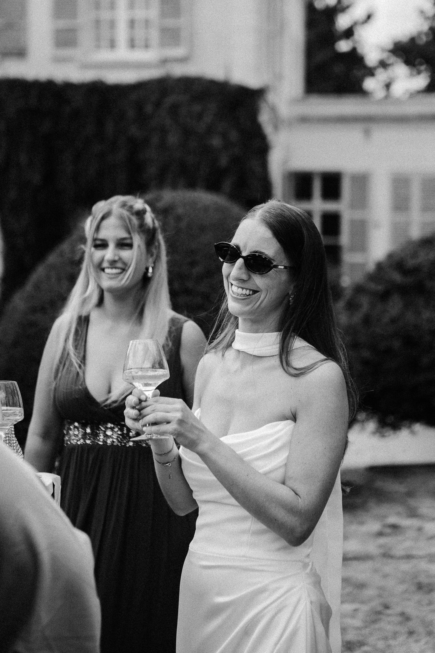 Black and white photo of smiling bride in white dress and sunglasses holding wine glass at outdoor wedding reception.