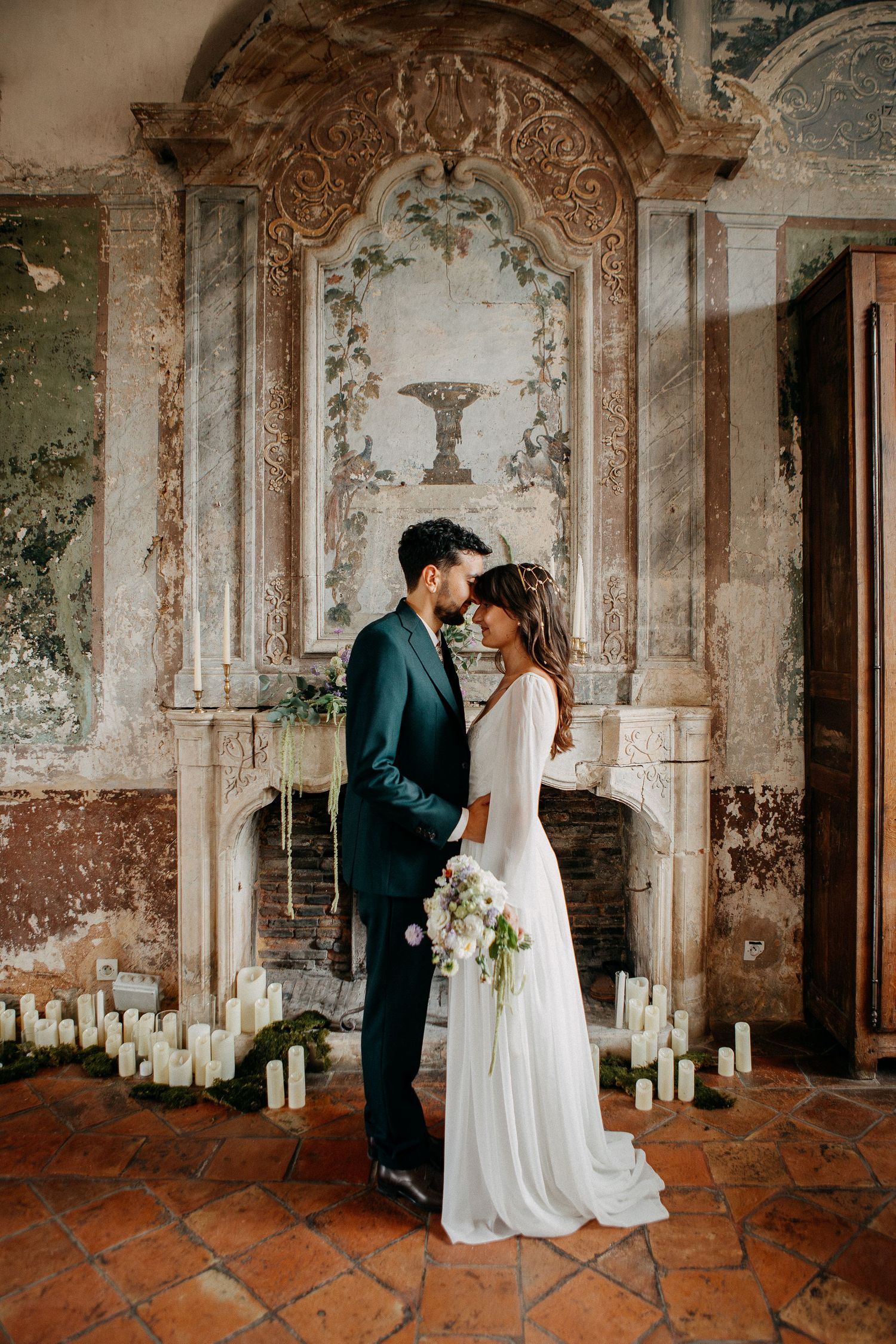 Bride and groom share an intimate moment before ornate fireplace surrounded by white candles in rustic venue.