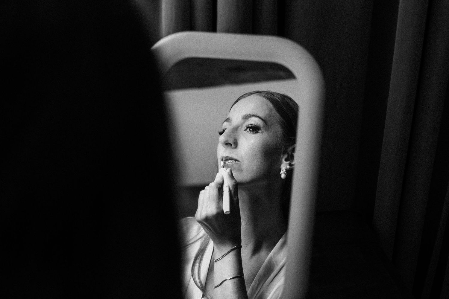 Bride applying makeup in mirror reflection, elegant black and white wedding preparation photo.