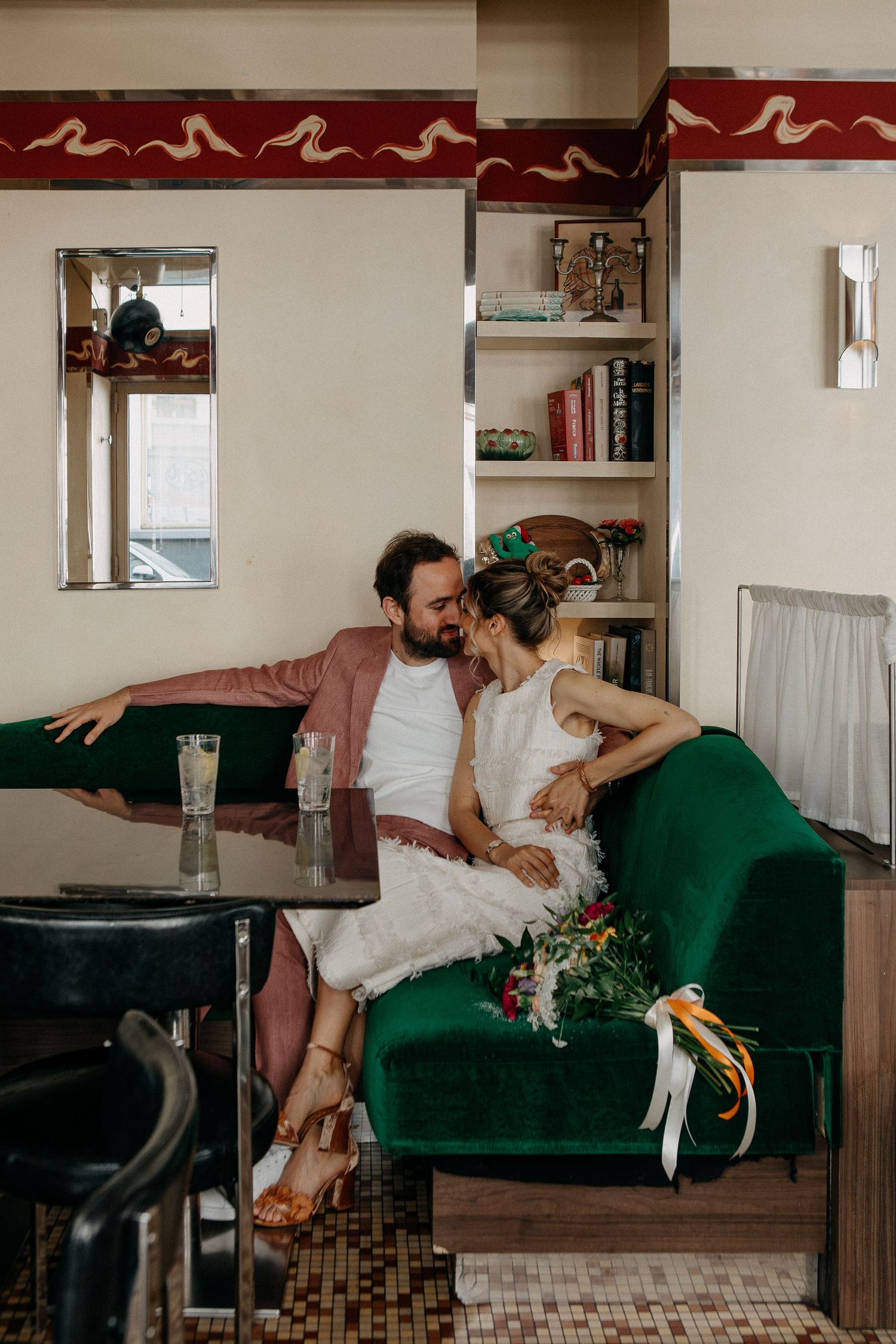 Couple sharing a romantic moment on a green velvet sofa in a vintage diner setting, bride holding a colorful bouquet.