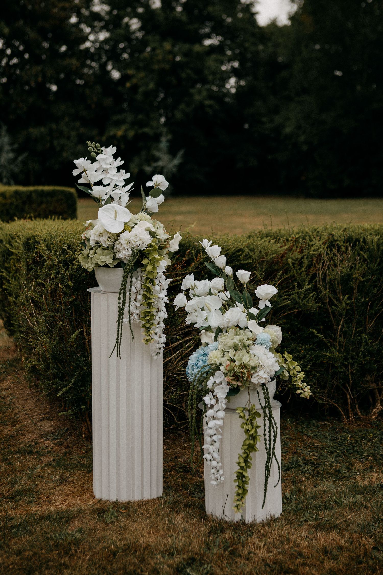 Two white fluted pedestals with elegant white and blue floral arrangements displayed outdoors on green grass.