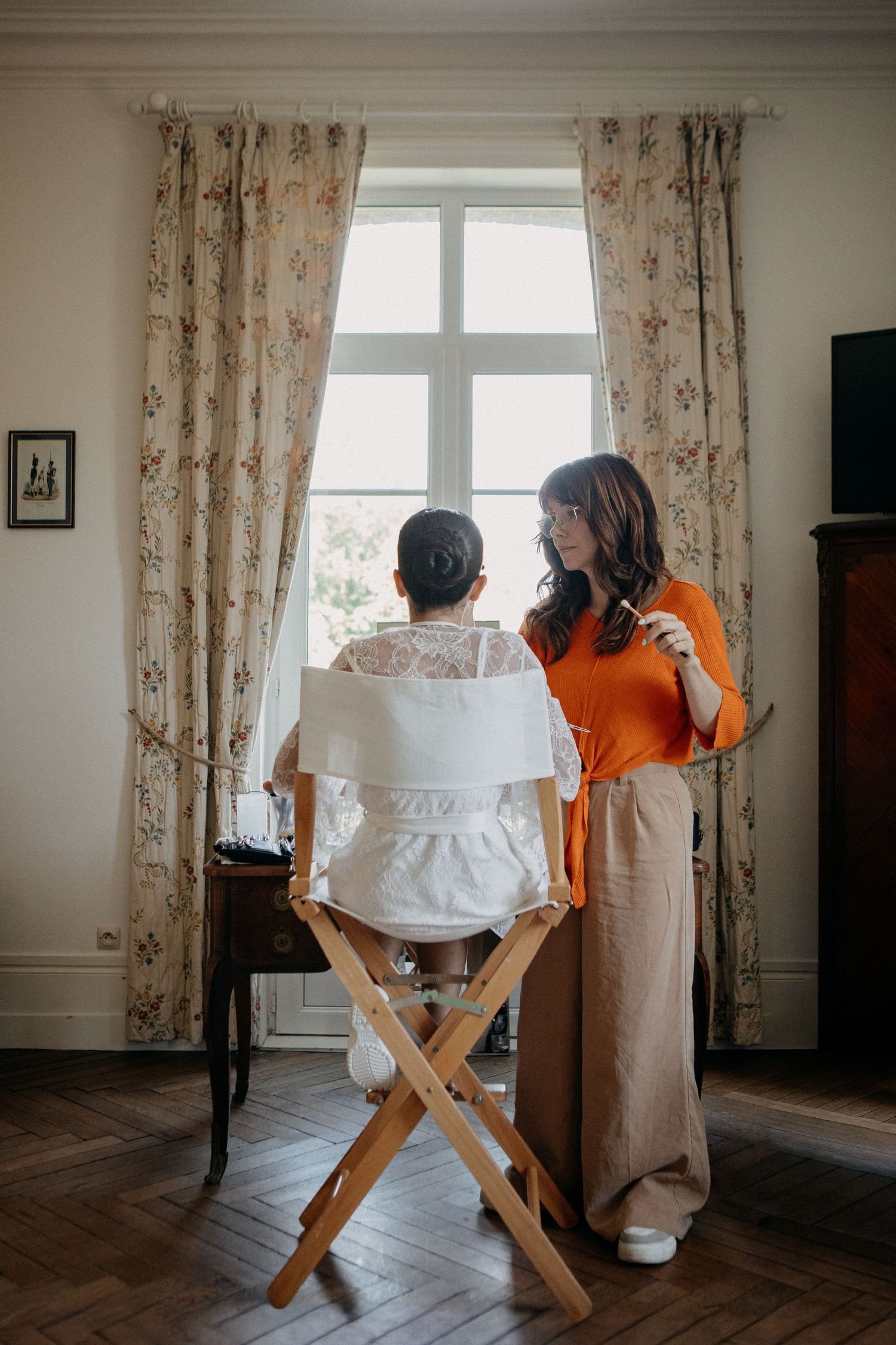 Bride in lace wedding dress sits by window while bridesmaid in orange top assists during bridal preparations.
