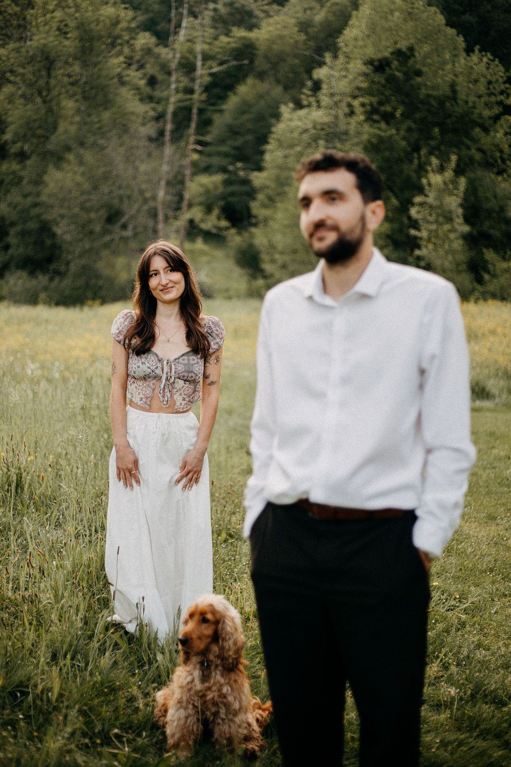 A smiling woman stands with a cocker spaniel while a bearded man poses in a green field.