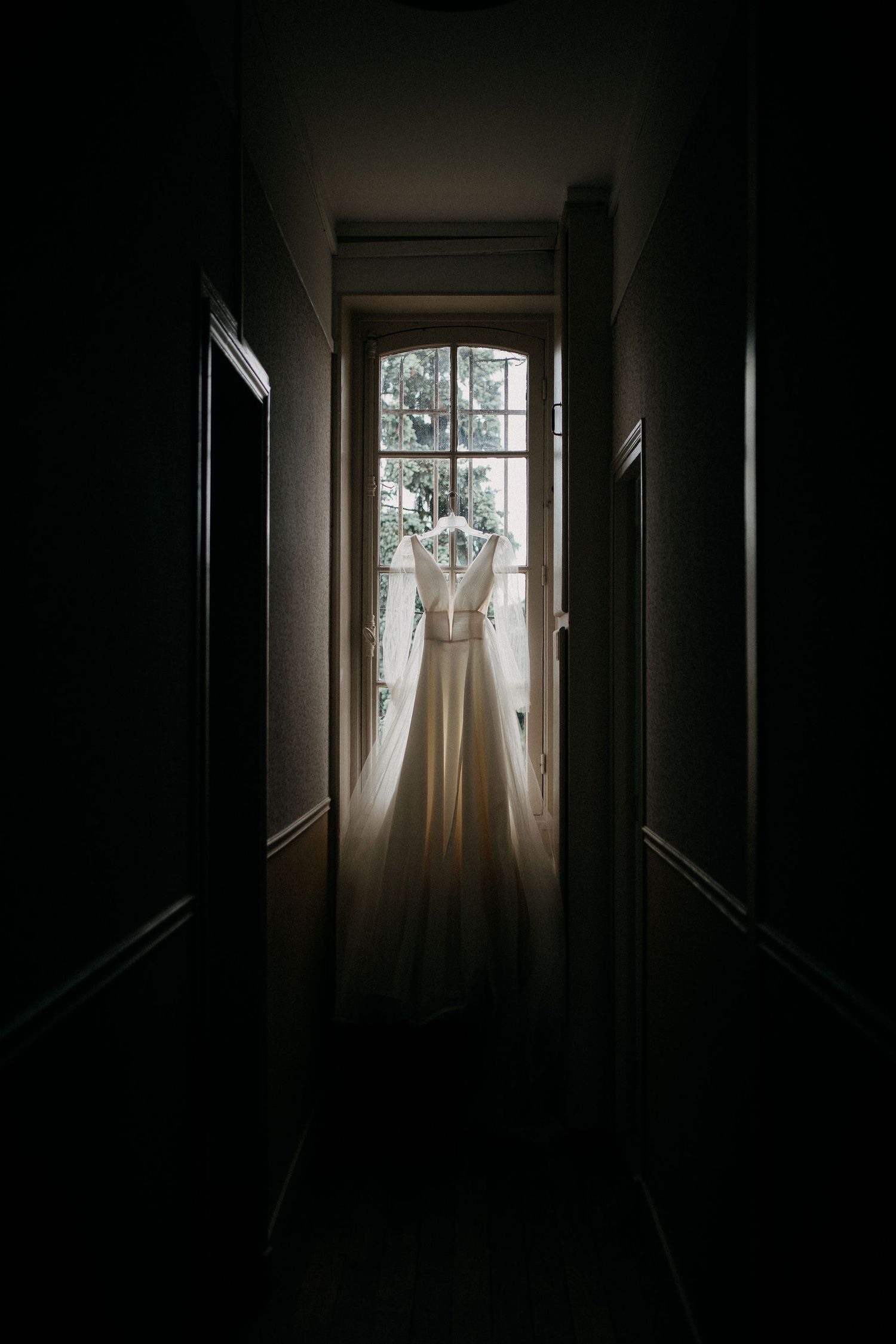 Ethereal bridal gown hanging in a dark hallway, backlit by a French window.