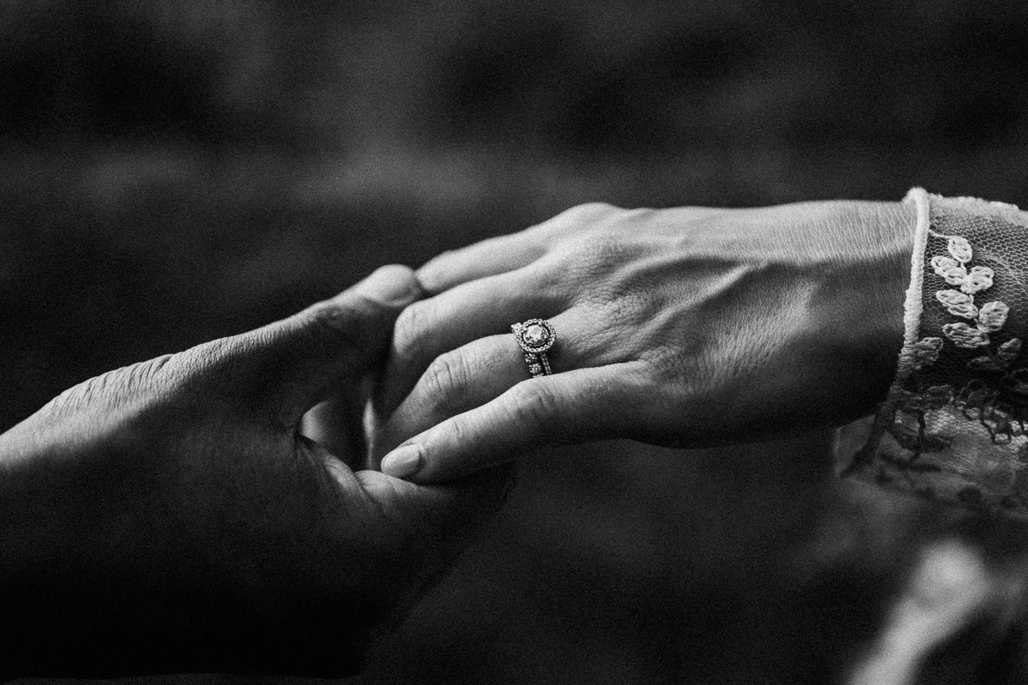 Black and white close-up of a couple holding hands, showing an elegant diamond engagement ring.