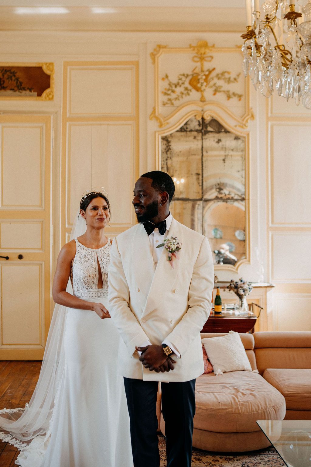 A bride in a lace gown stands behind her groom in a cream tuxedo for a first look.