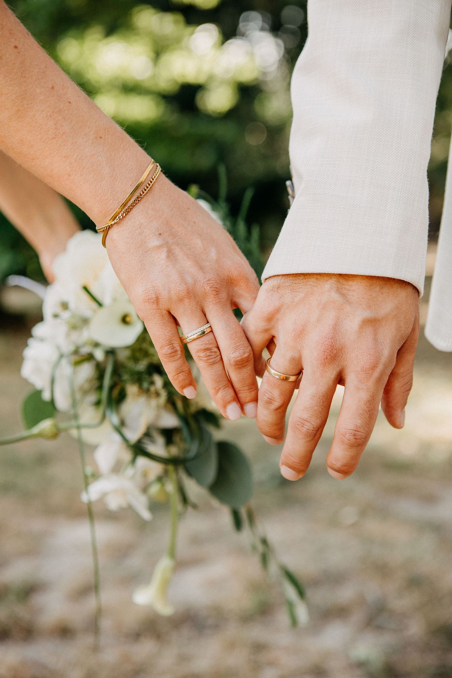Newlywed couple holding hands displaying wedding rings with bridal bouquet of white flowers and eucalyptus.