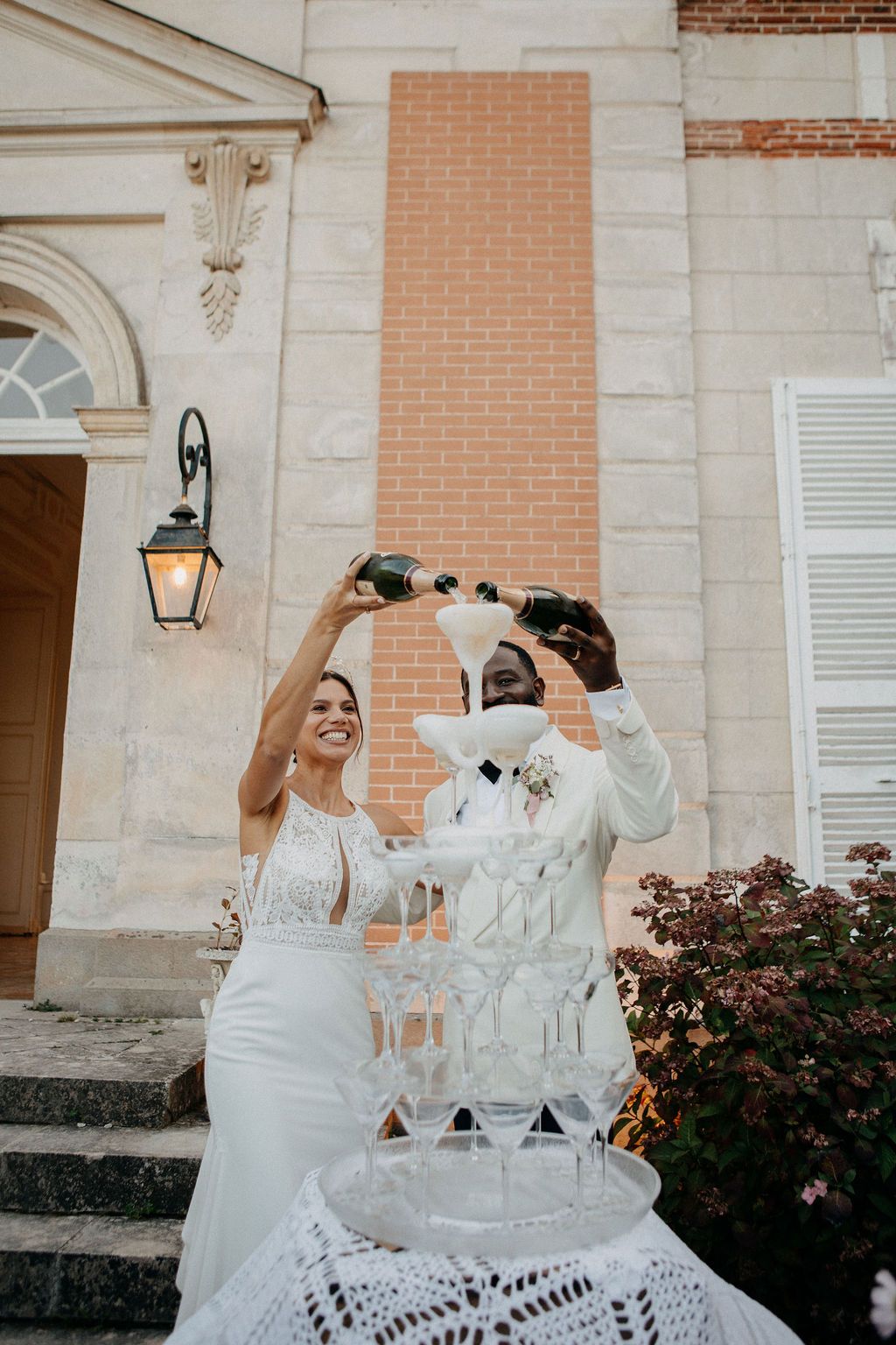 Bride and groom joyfully pouring champagne into a tiered champagne tower outside a French chateau.