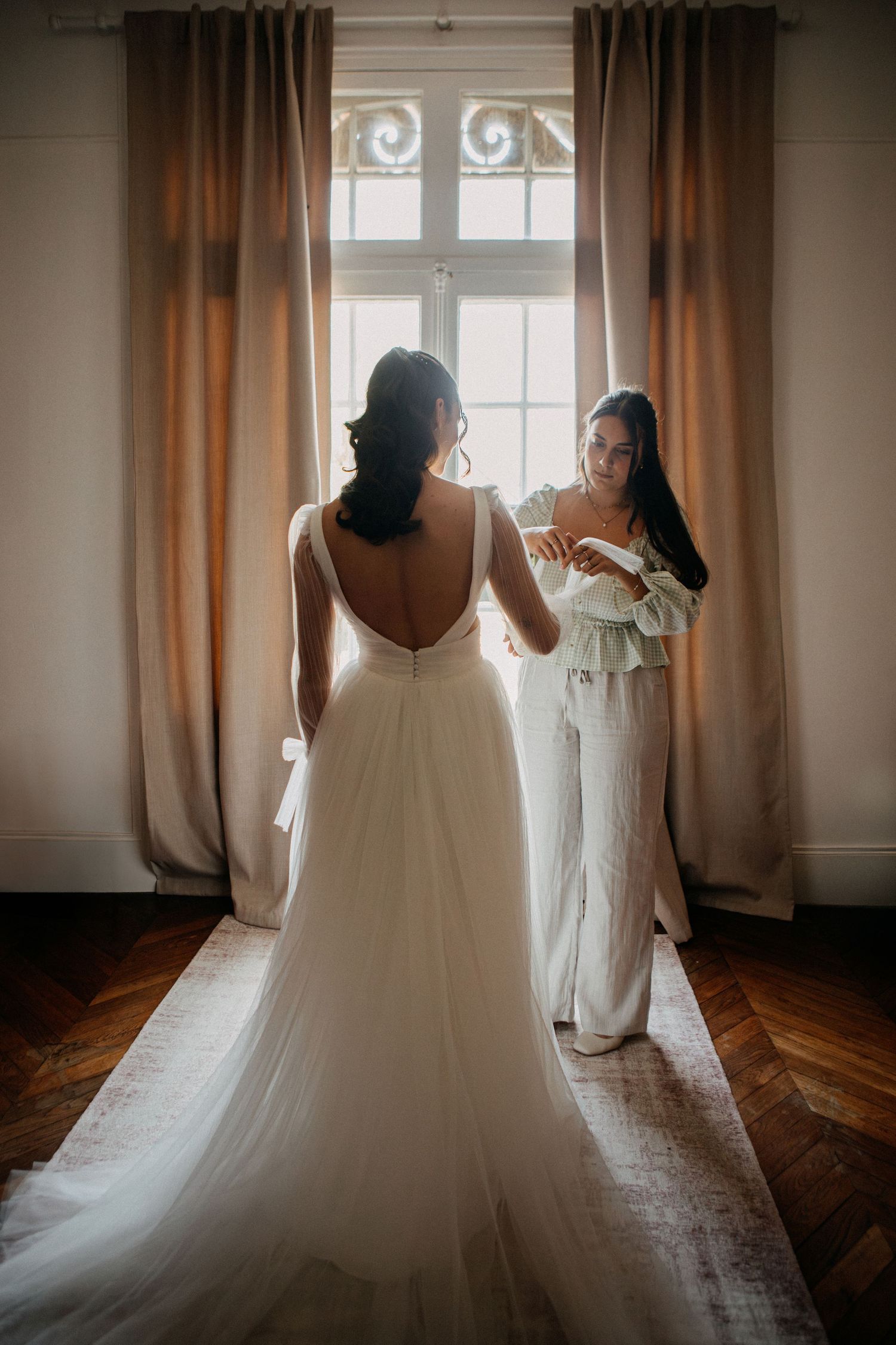 Bride in elegant backless white wedding gown being helped by bridesmaid near sunlit window.
