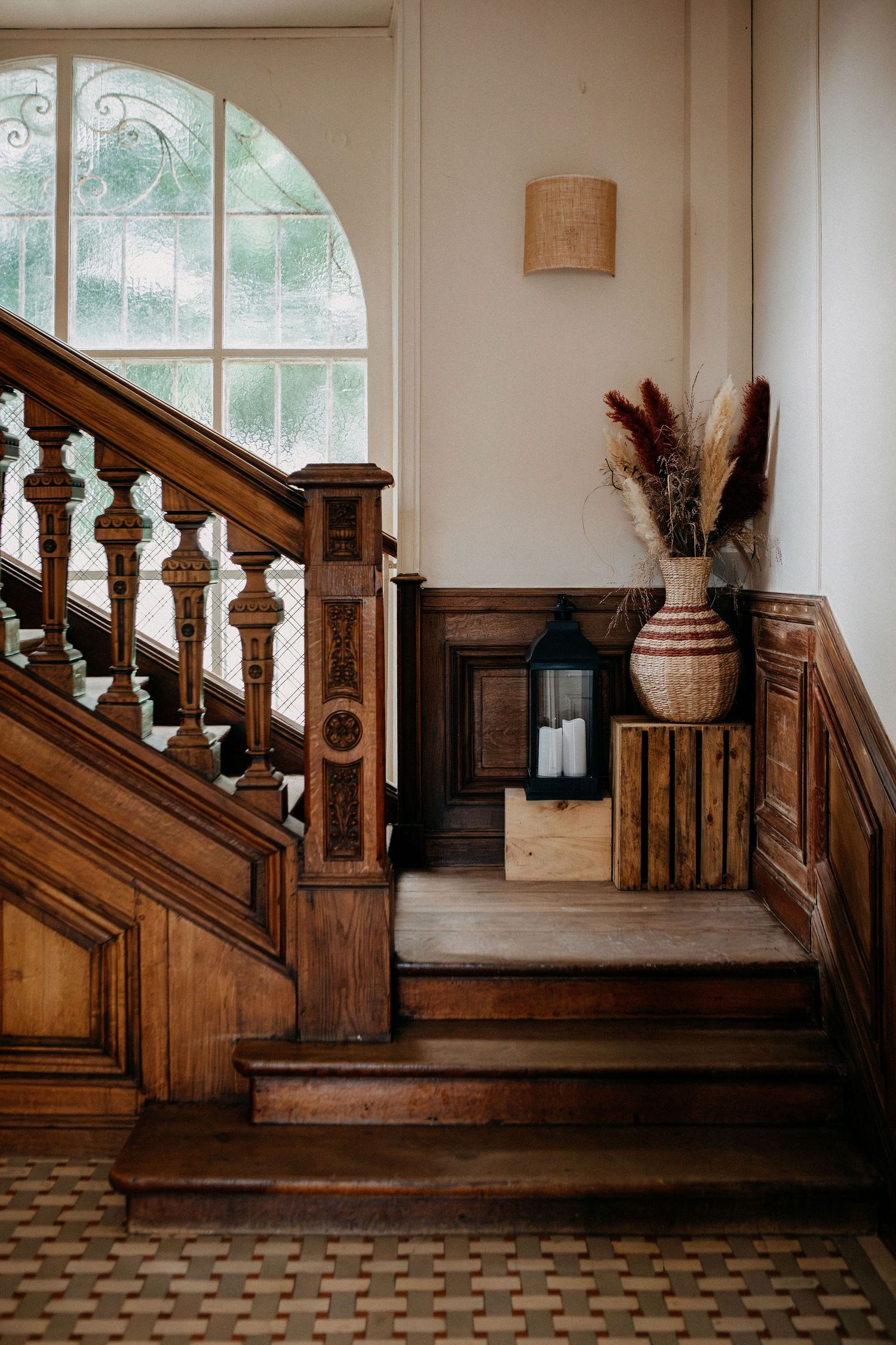 Ornate wooden staircase with carved banister in a classic French manor with arched stained glass window.