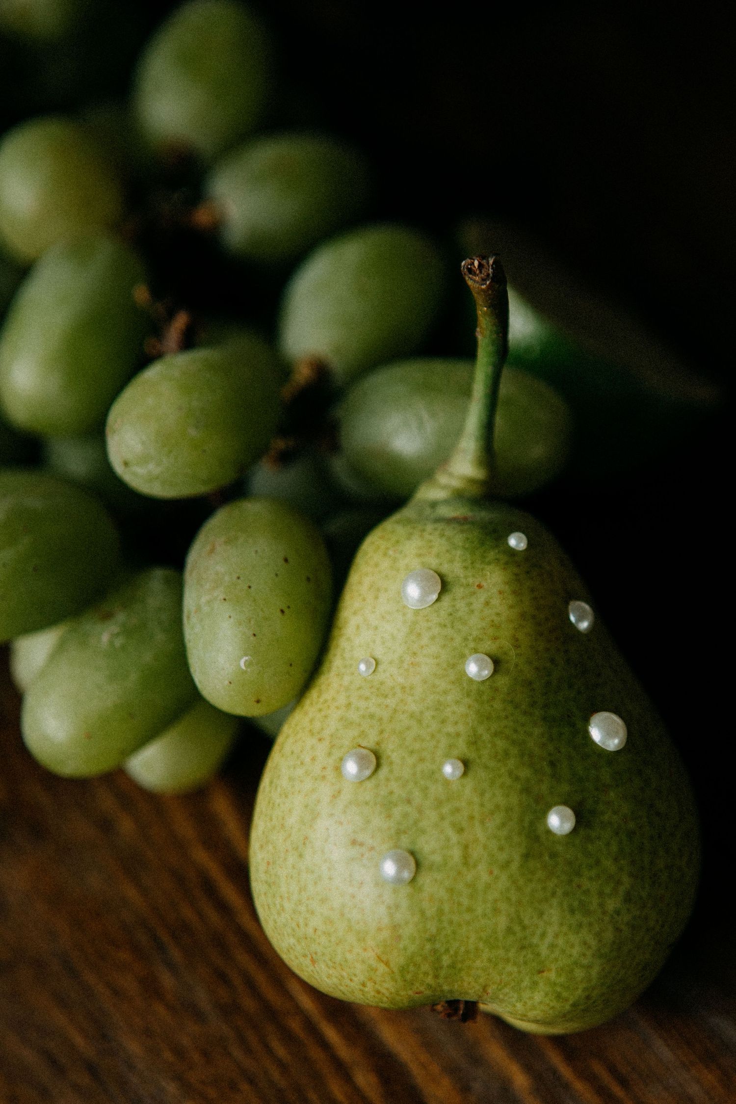 Green pear decorated with pearl beads beside a bunch of fresh green grapes on a dark wooden surface.