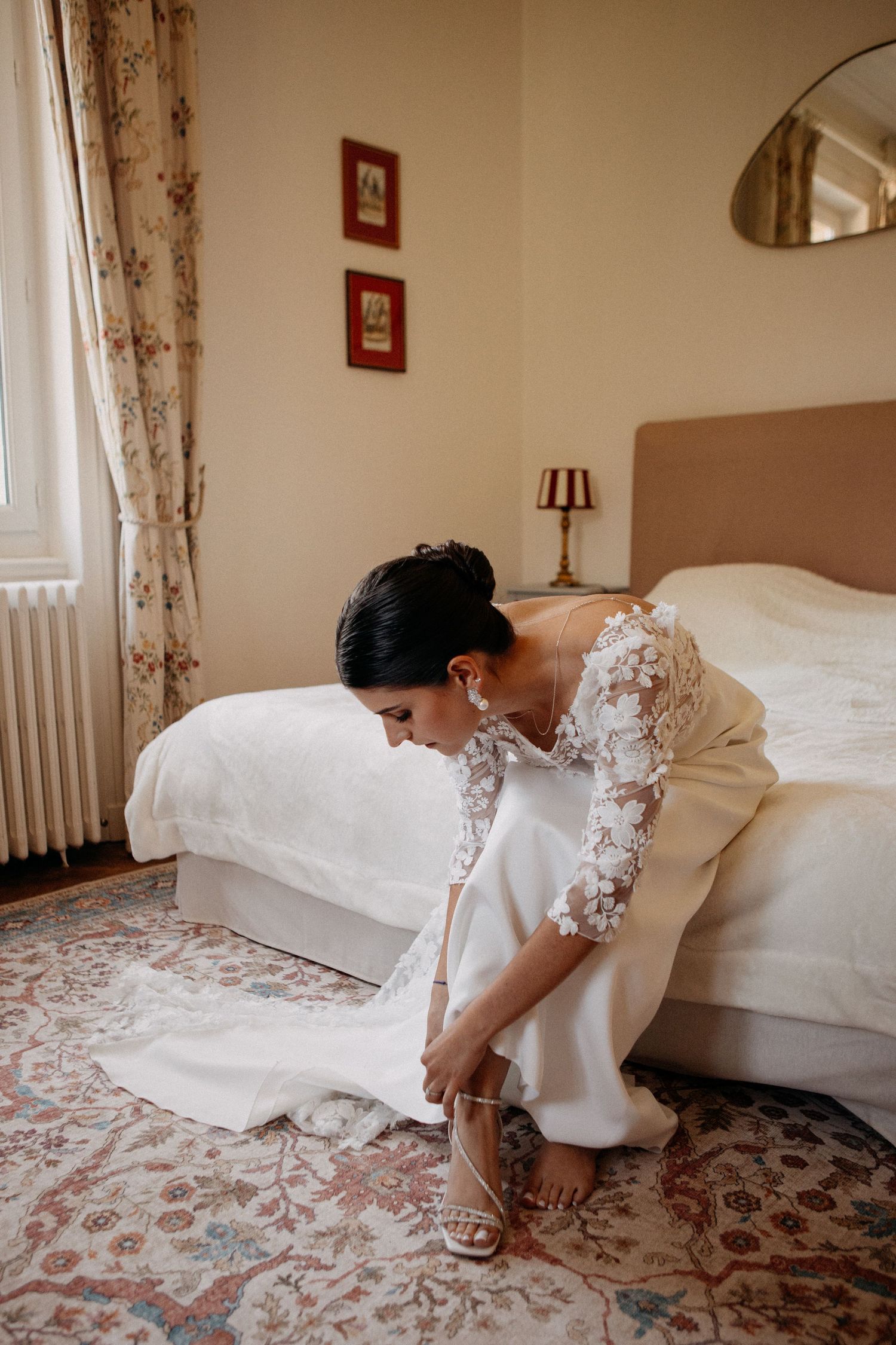 Bride in long-sleeve lace wedding gown sits on bed edge fastening strappy heeled sandals in bedroom.