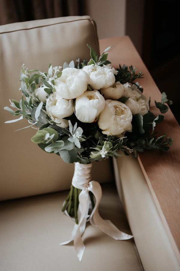 Elegant bridal bouquet of white peonies and greenery tied with ribbon on a chair.
