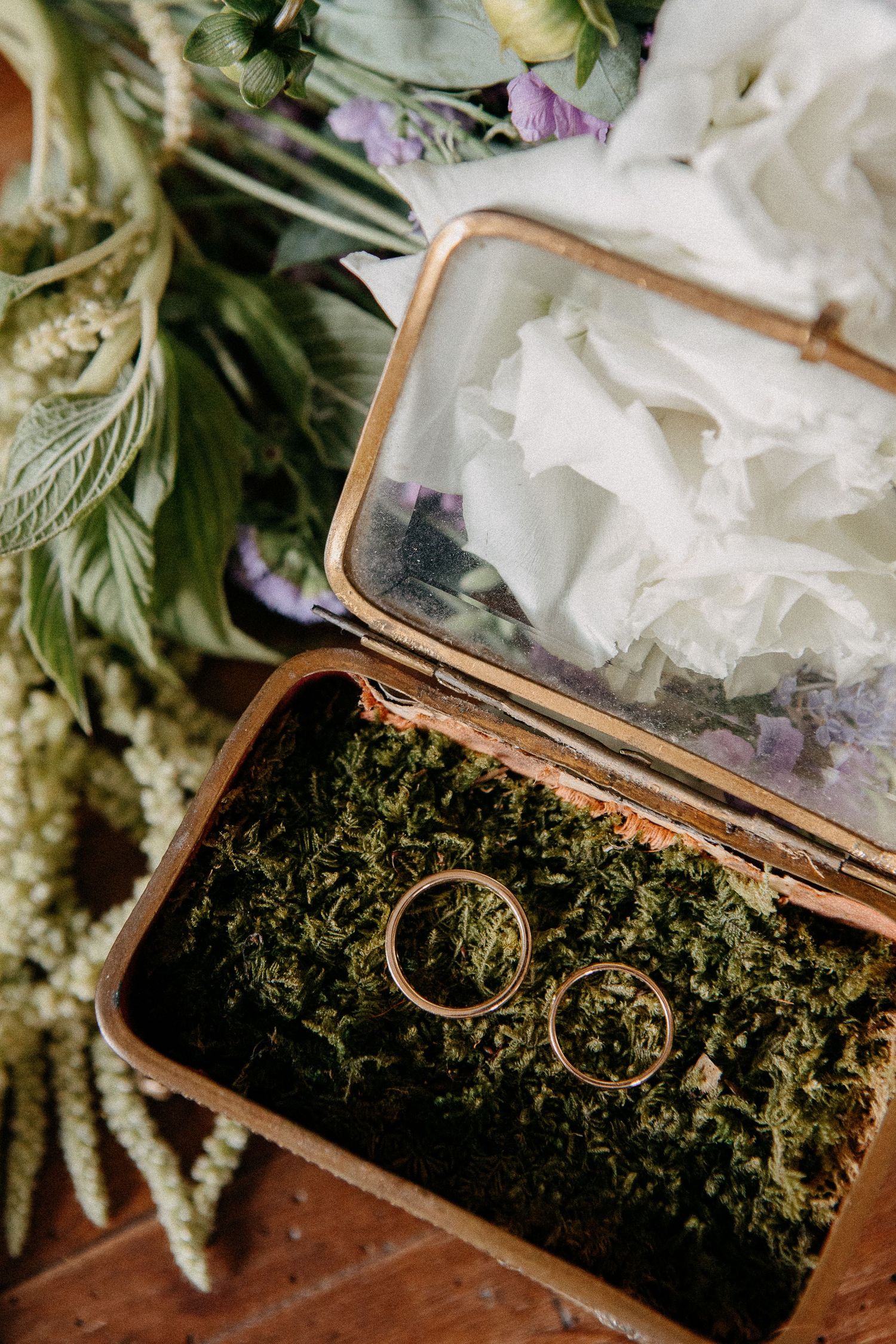 Two gold wedding rings rest on moss inside a vintage brass glass-top box, surrounded by a lush floral bouquet.