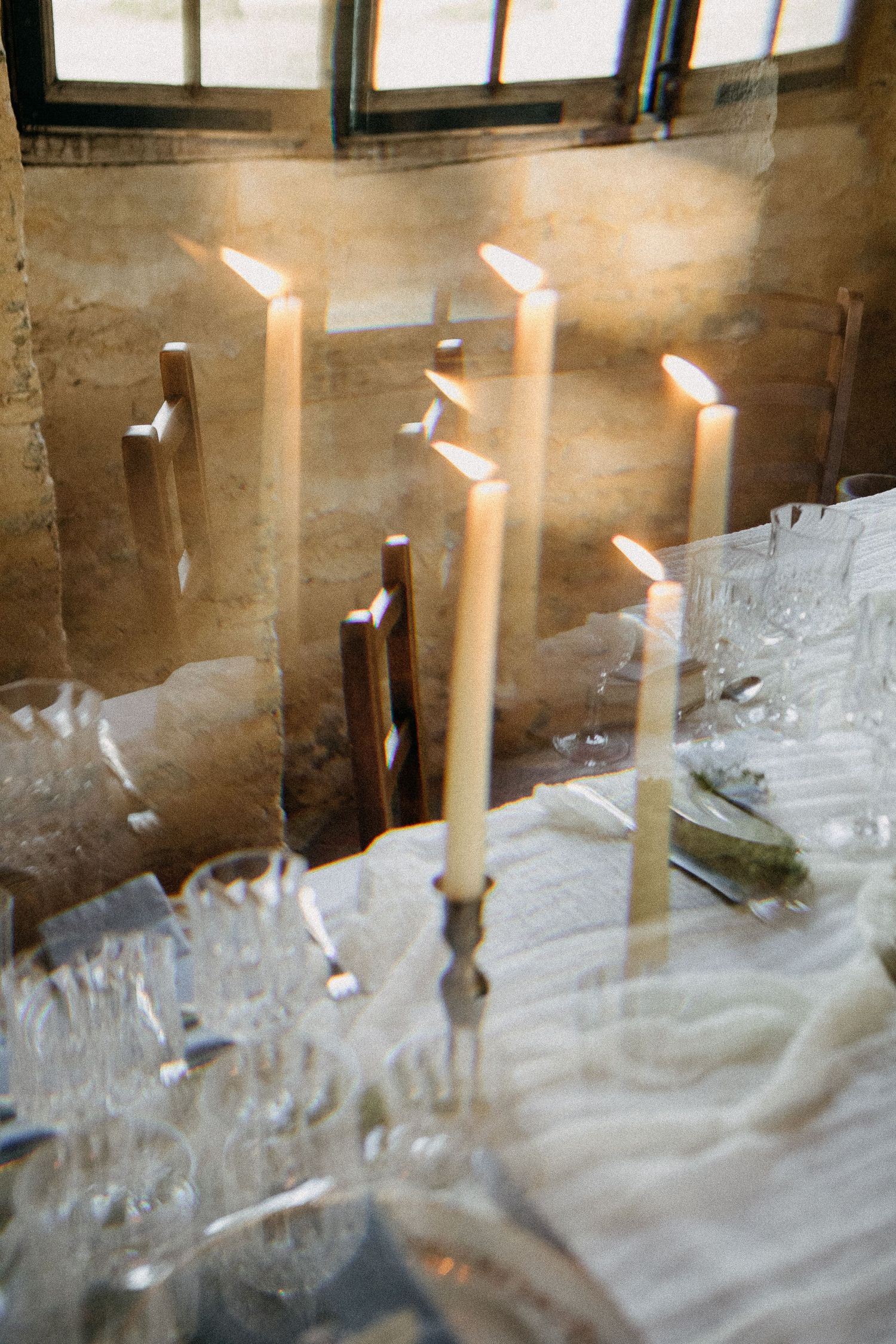 Elegant candlelit dining table with tall white candles, crystal glasses, and soft natural light in a rustic stone venue.