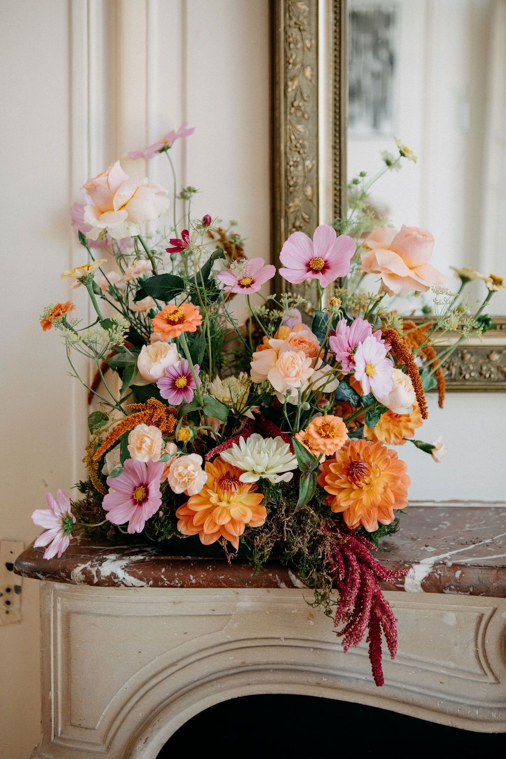Lush bridal bouquet with orange dahlias, pink cosmos, and peach roses displayed on a marble mantelpiece.