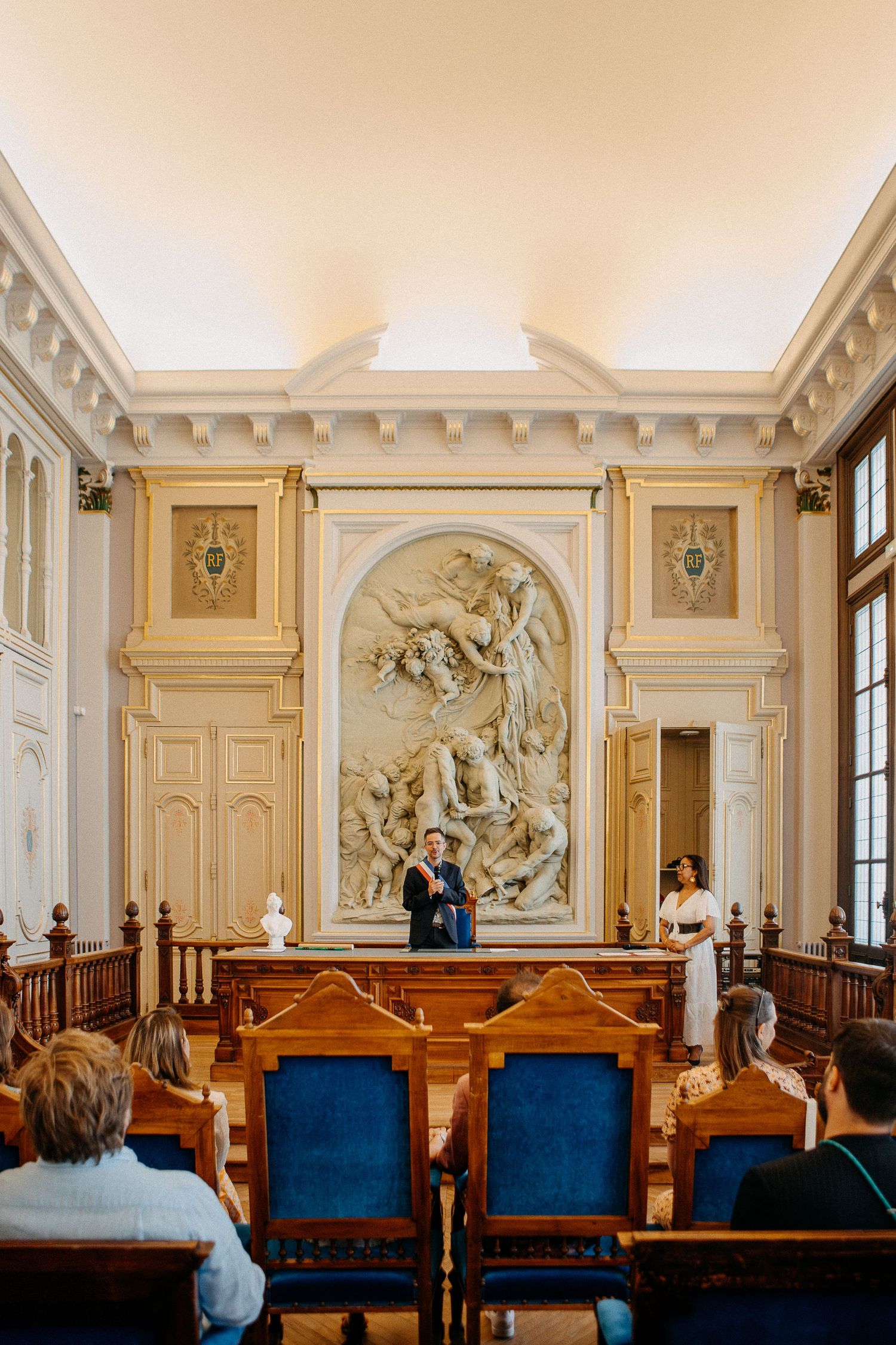 Elegant wedding ceremony in ornate hall with baroque relief, blue velvet chairs, and couple at wooden altar.
