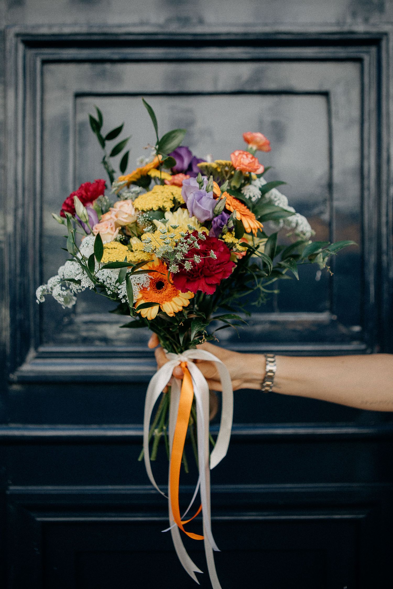 A hand holding a vibrant mixed flower bouquet with ribbons against a dark door.