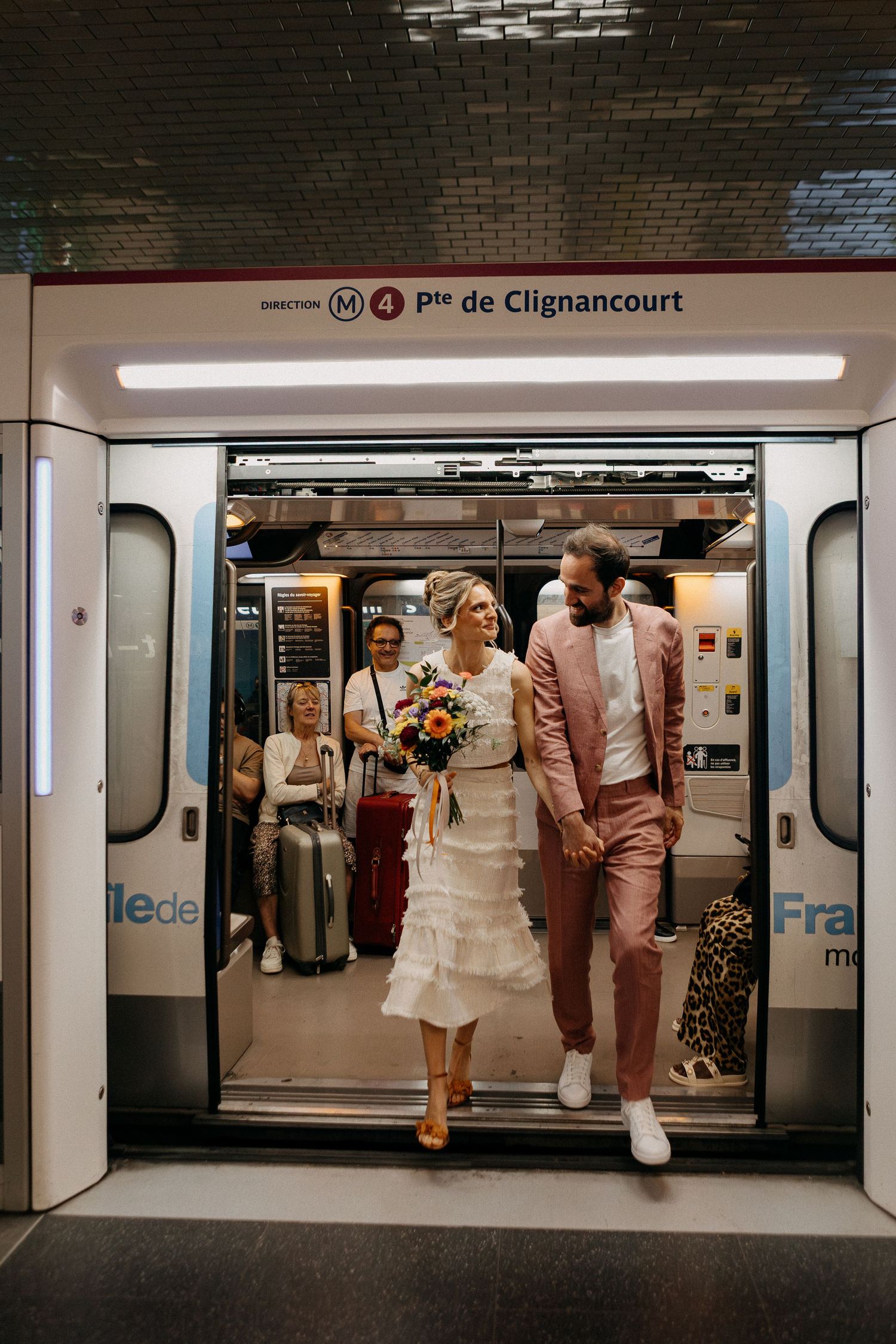 Bride and groom exit Paris Metro Line 4 train in wedding attire, bride holding colorful bouquet at Porte de Clignancourt.