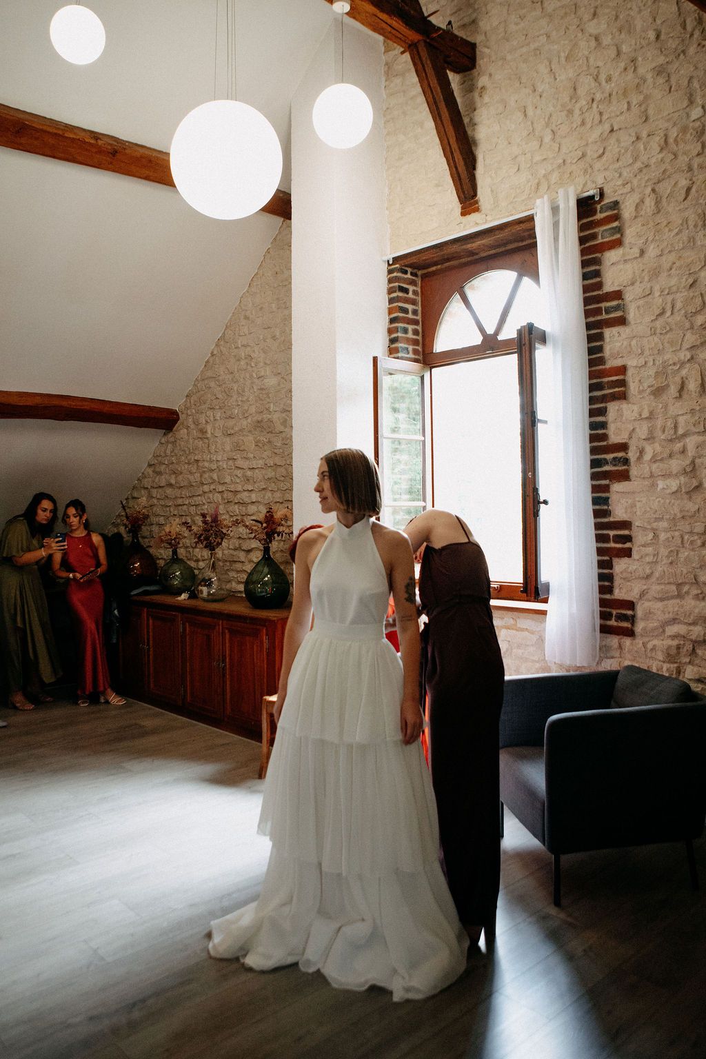 Bridesmaid in burgundy dress fastening bride's tiered white wedding gown in rustic stone venue.