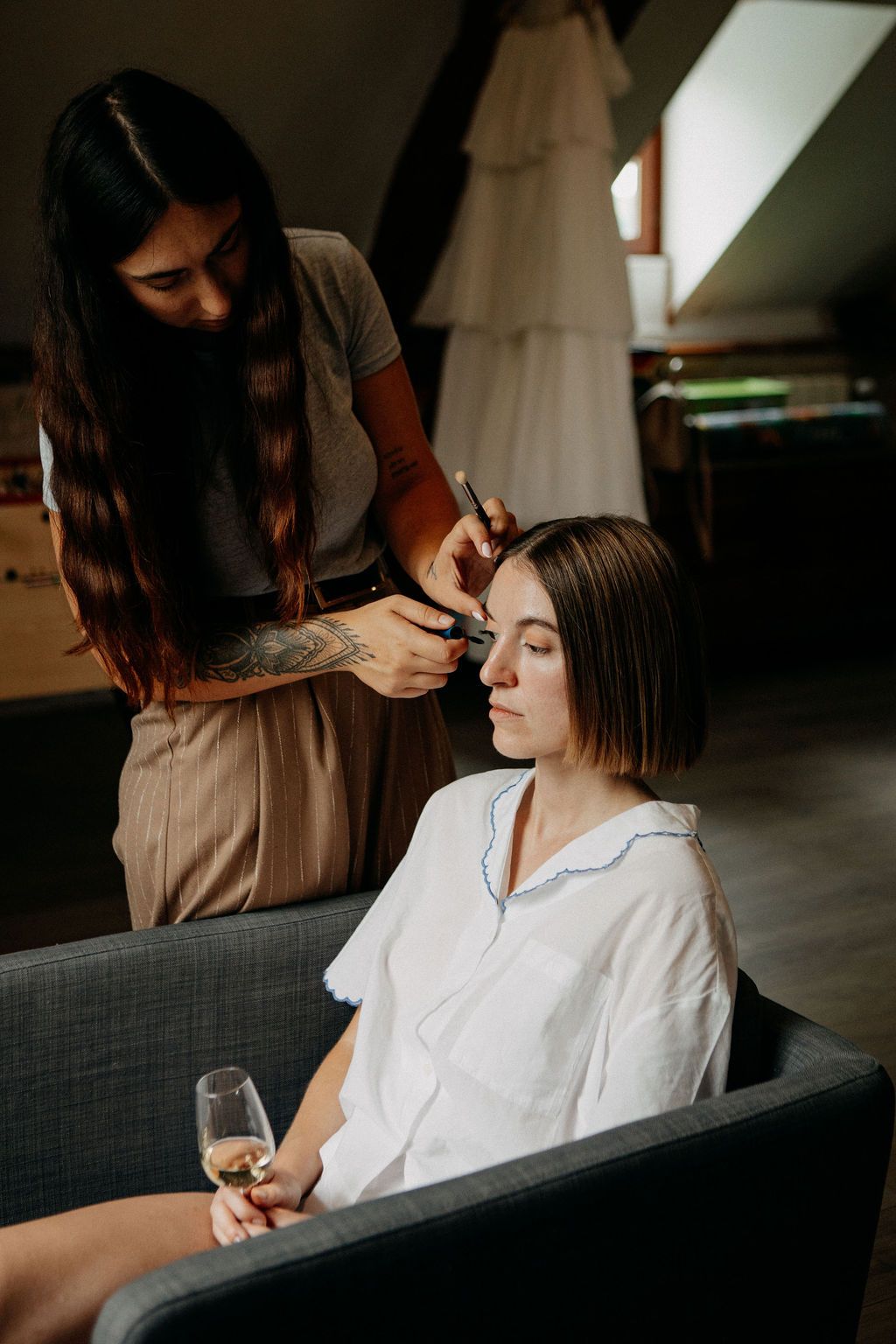 Makeup artist applying eyeshadow to seated bride holding champagne glass in dressing room.