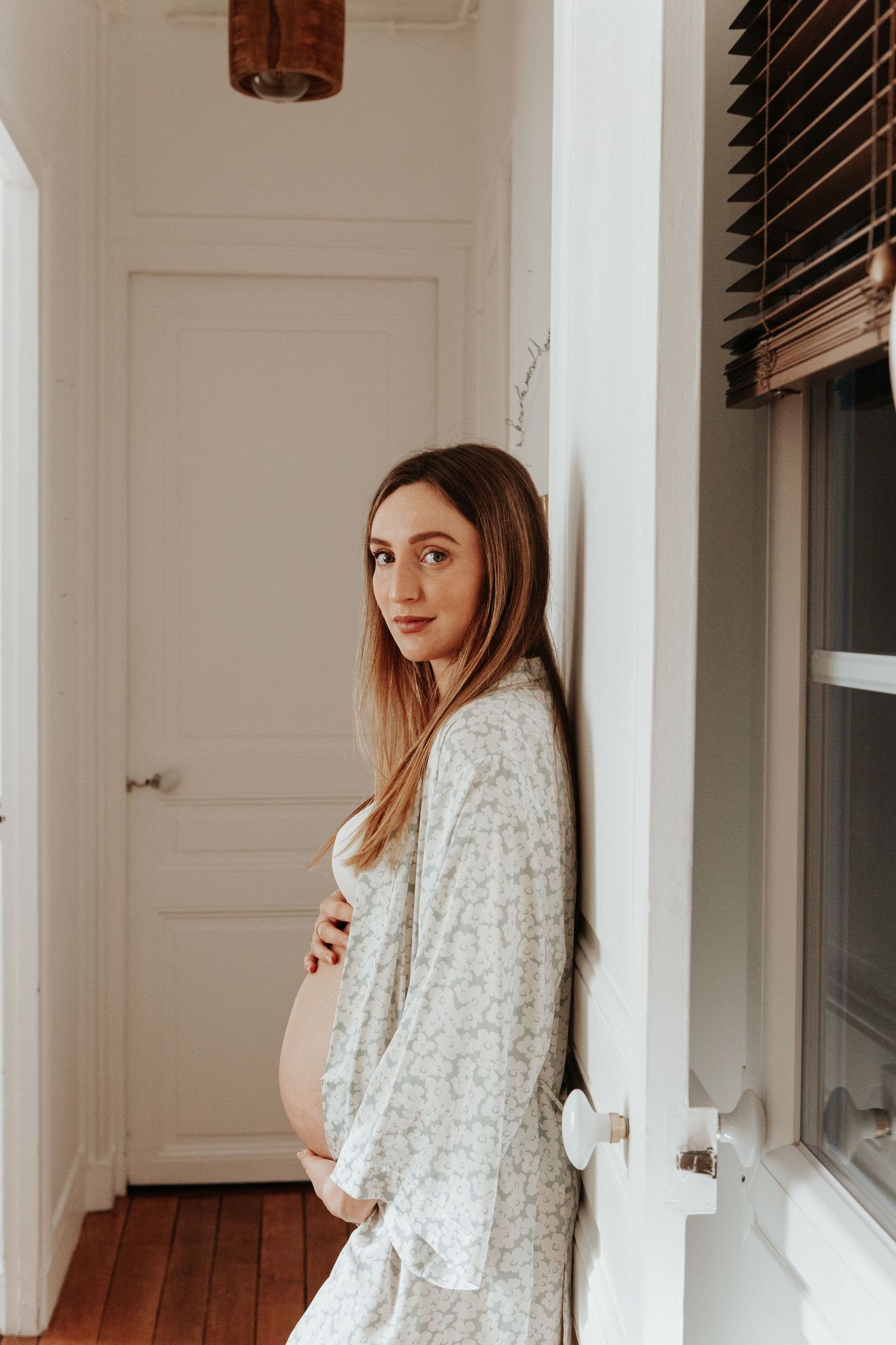 Pregnant woman in floral robe holding belly while leaning against wall by window.