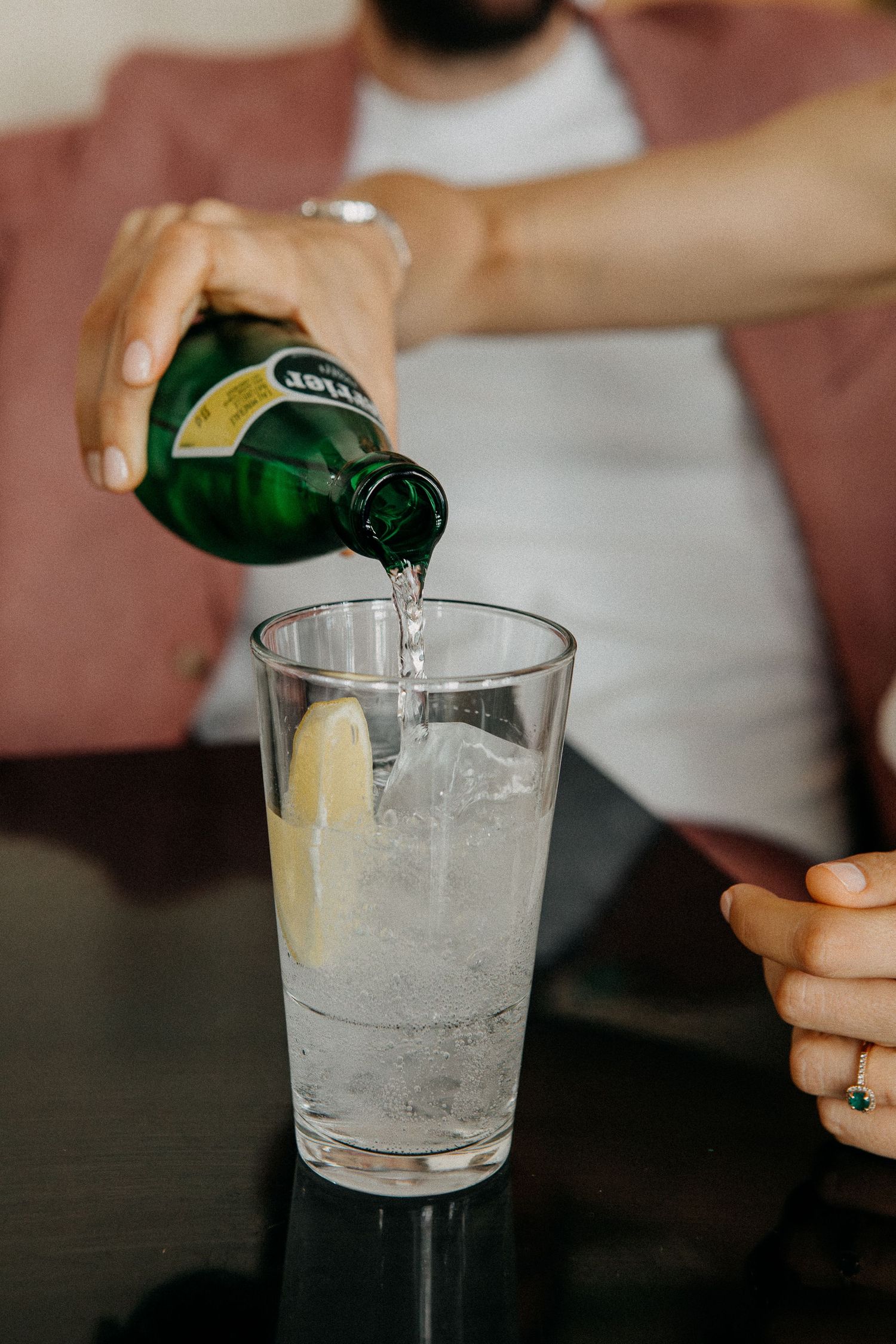 Person pouring sparkling water from green bottle into glass with ice and lemon slice on dark table.