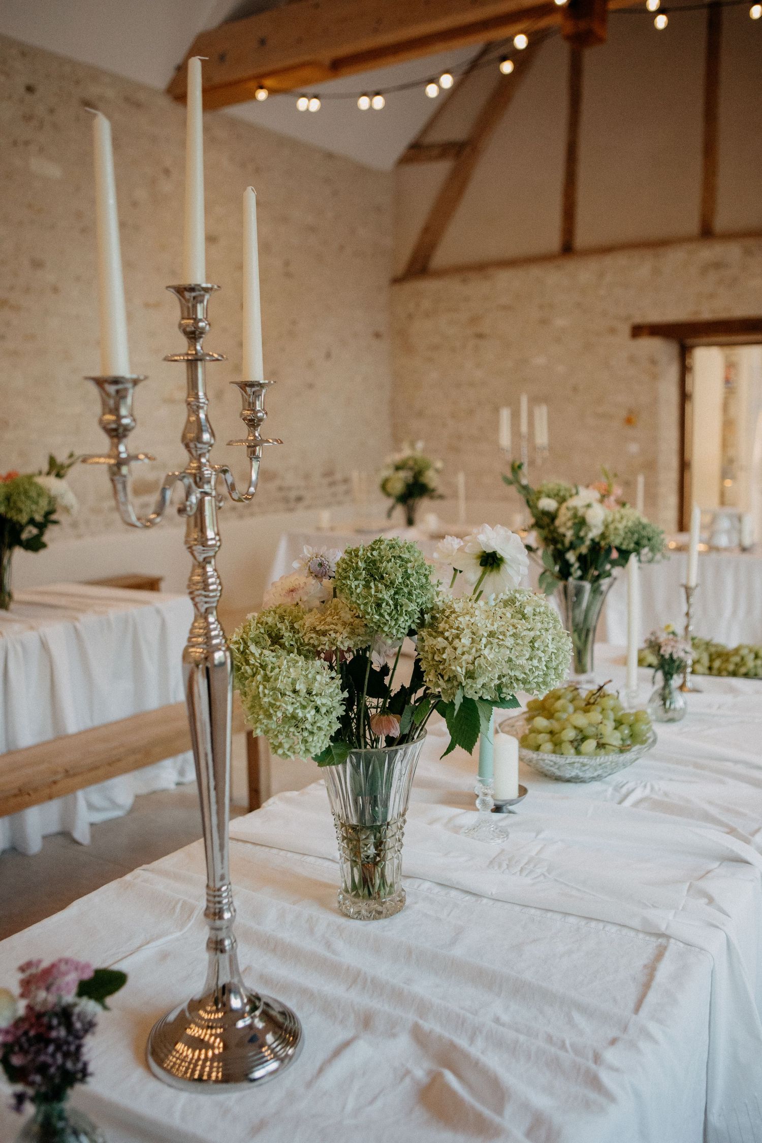 Elegant wedding table with silver candelabra, white hydrangea centerpieces, and string lights in barn venue.