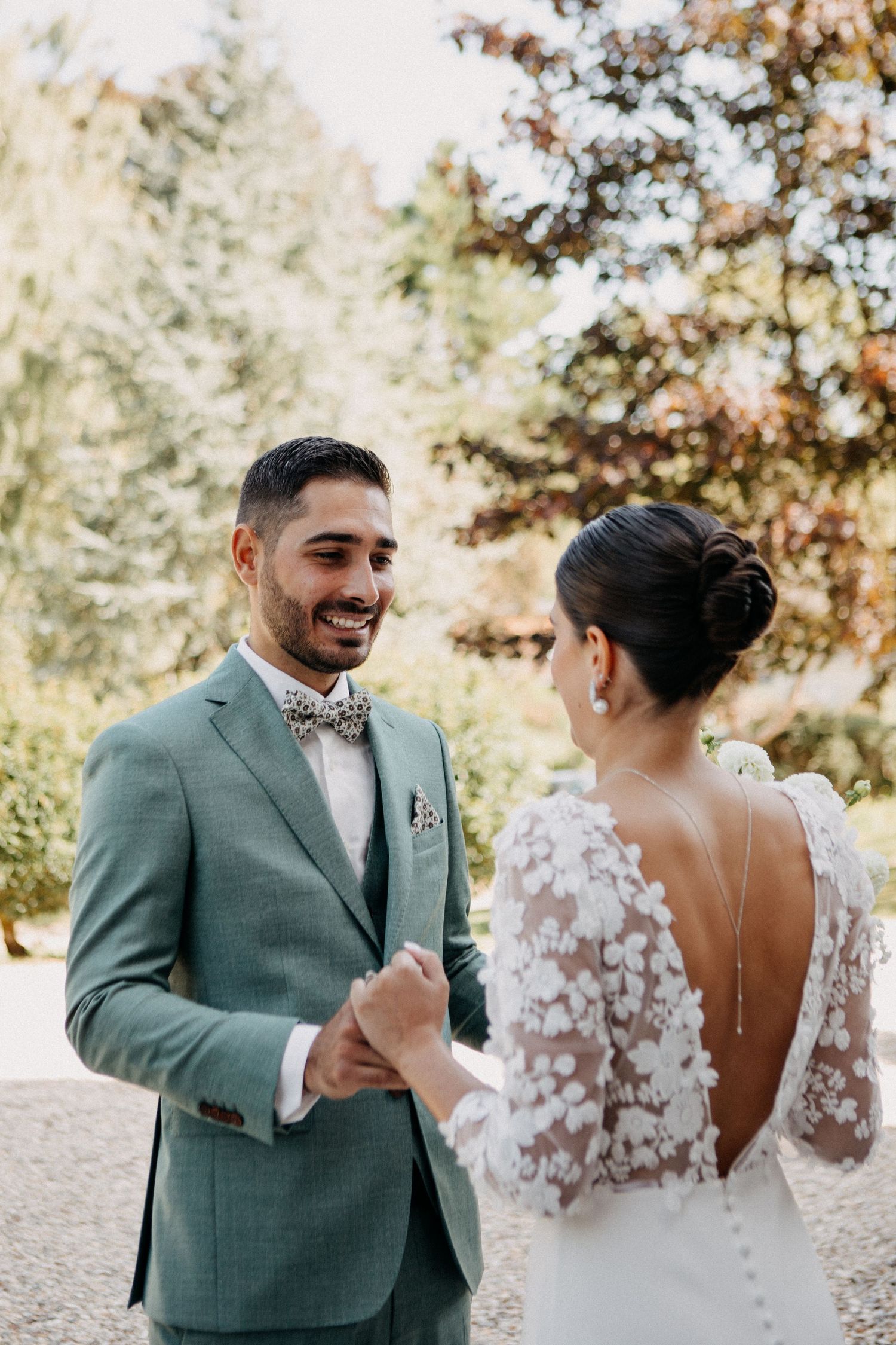 Smiling groom in sage suit holds bride's hands as she faces him in floral lace backless wedding gown outdoors.