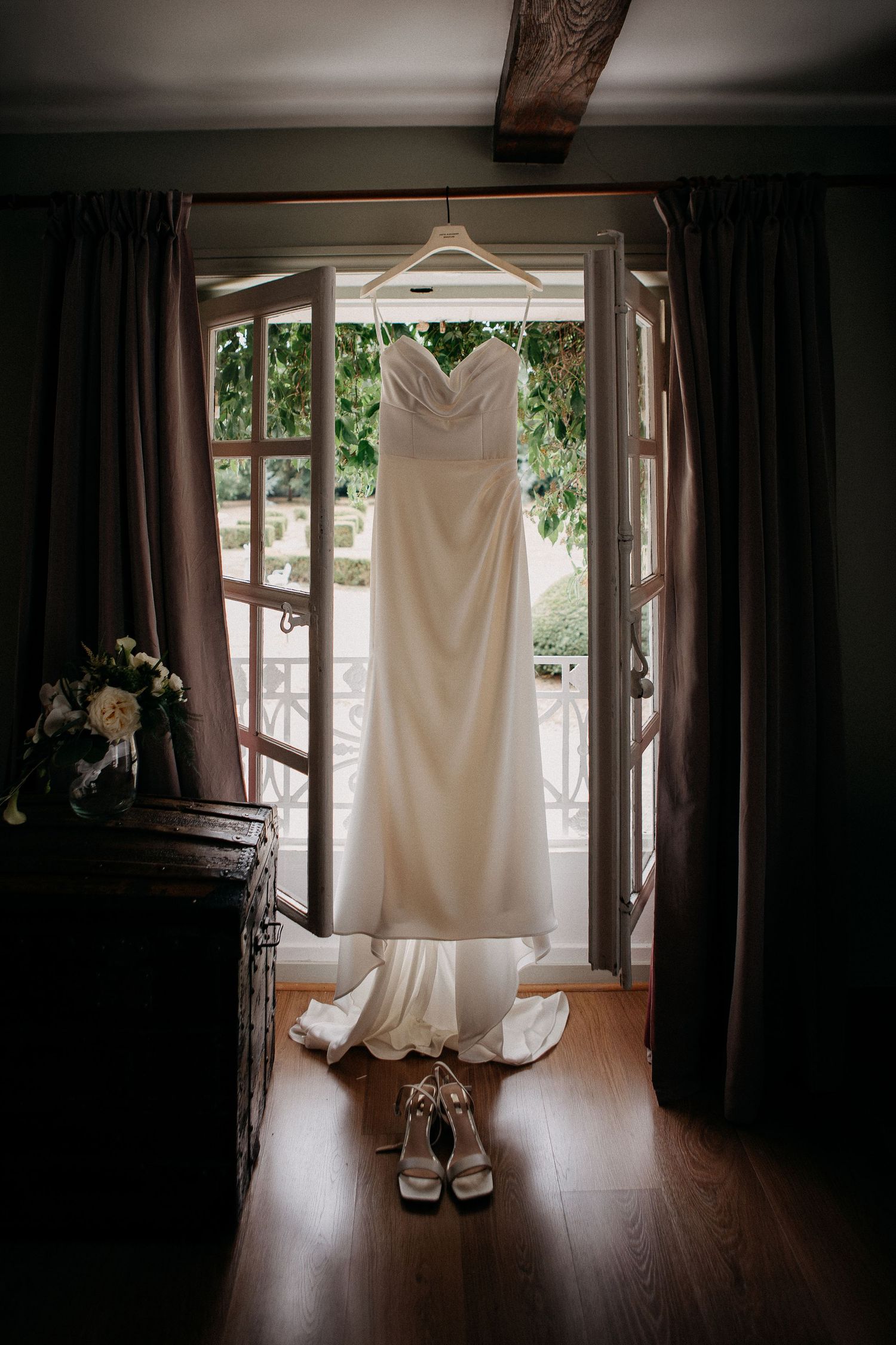 Elegant white wedding dress hanging in doorway with bridal heels below, soft natural light, dark wood floors.