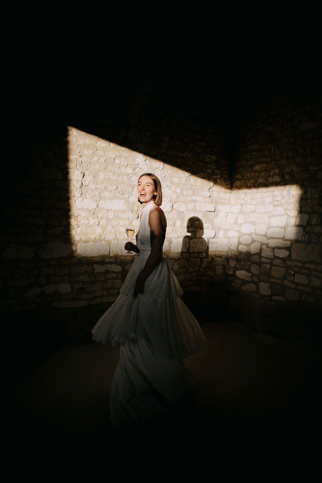 Radiant bride in tiered gown twirling with champagne in dramatic stone wall light beam.