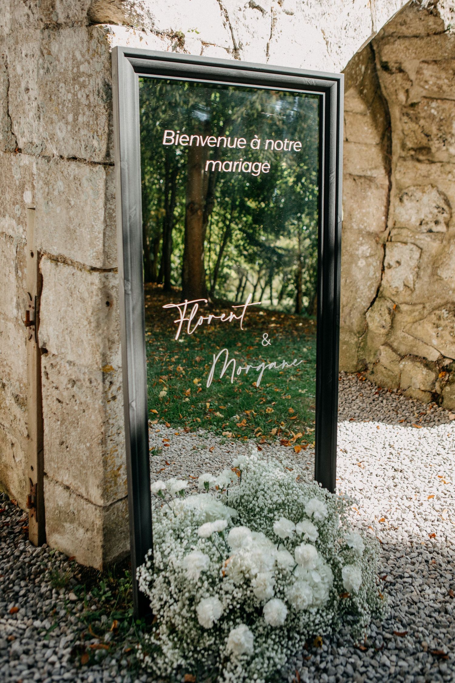 Wedding welcome mirror sign reading Bienvenue a notre mariage Florent and Morgane with white floral arrangement.