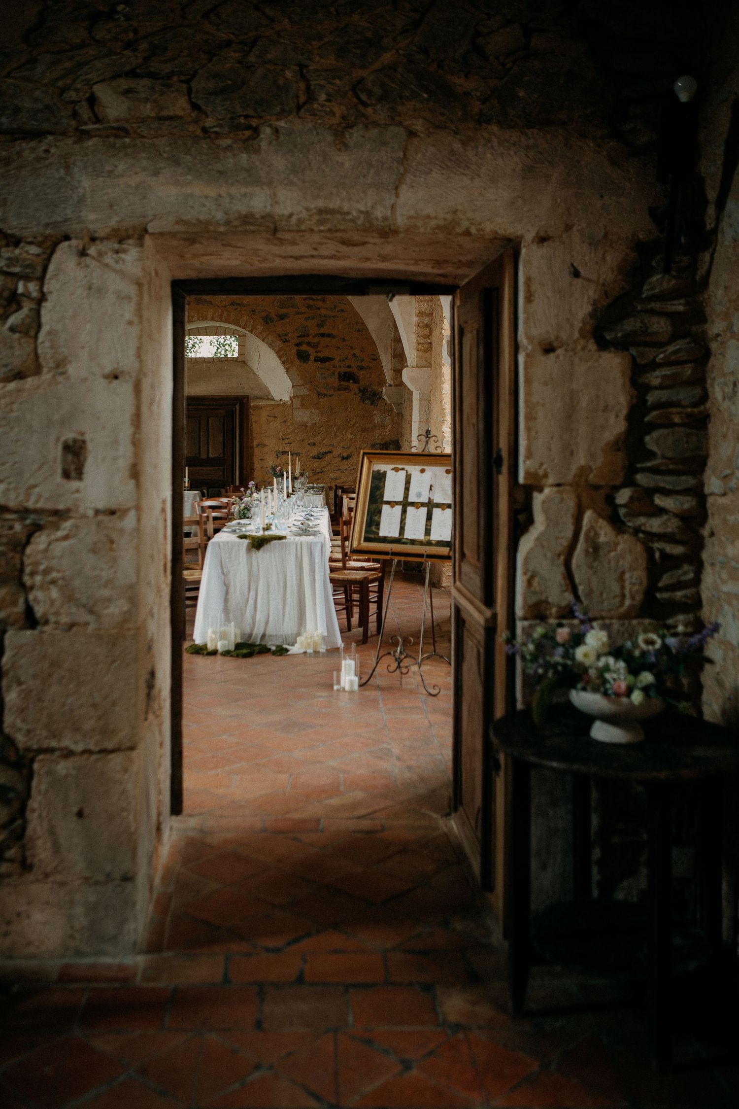 Rustic stone venue with elegant white-draped wedding table and floral arrangements visible through an arched doorway.