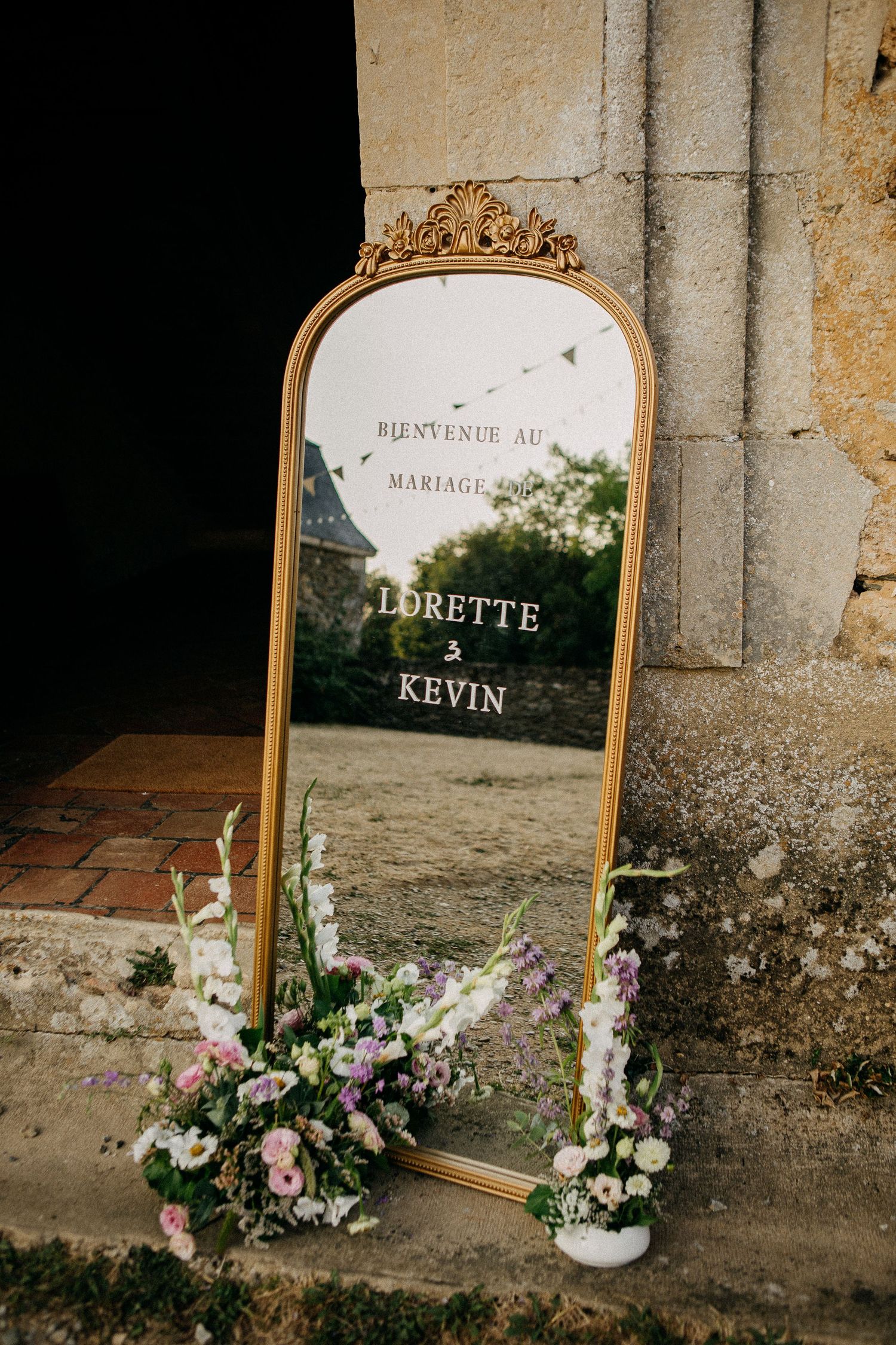 Gold ornate mirror wedding welcome sign for Lorette and Kevin, adorned with fresh floral arrangements at base.