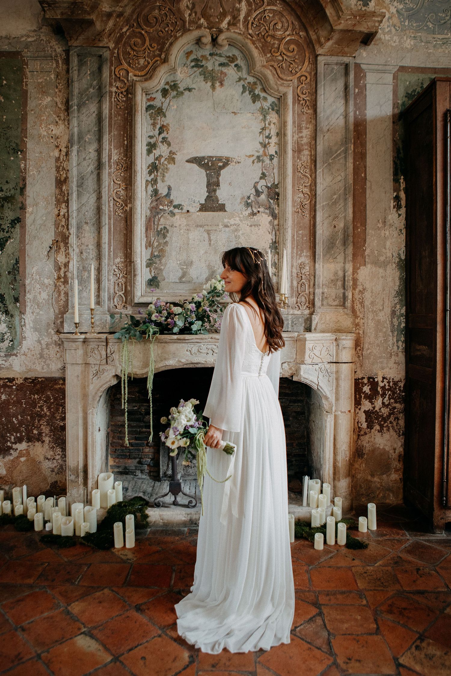 Bride in flowing white gown stands before ornate fireplace with candles and floral arrangements in elegant historic venue.