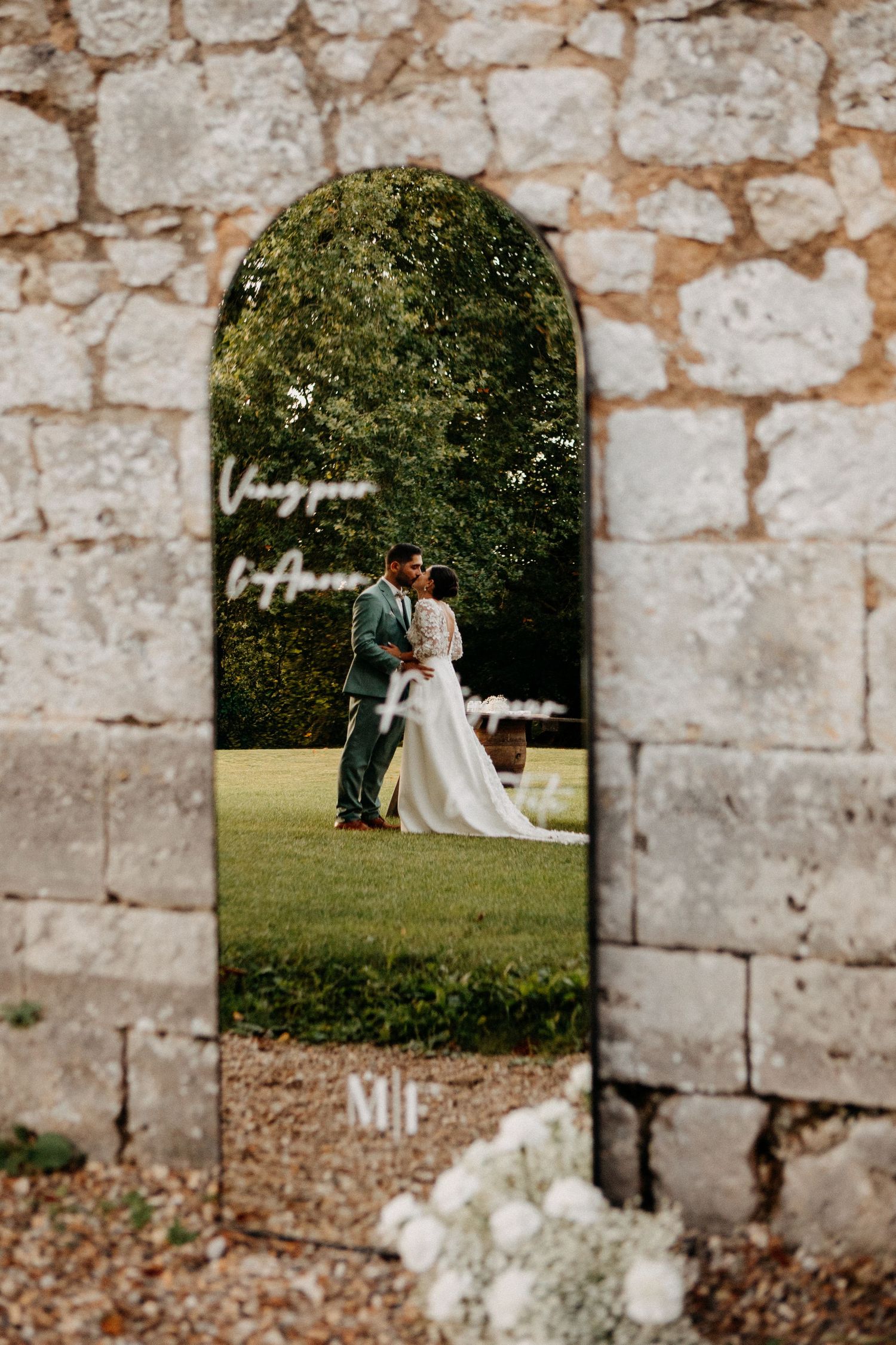 Couple sharing romantic kiss reflected in arched mirror at outdoor garden wedding venue.