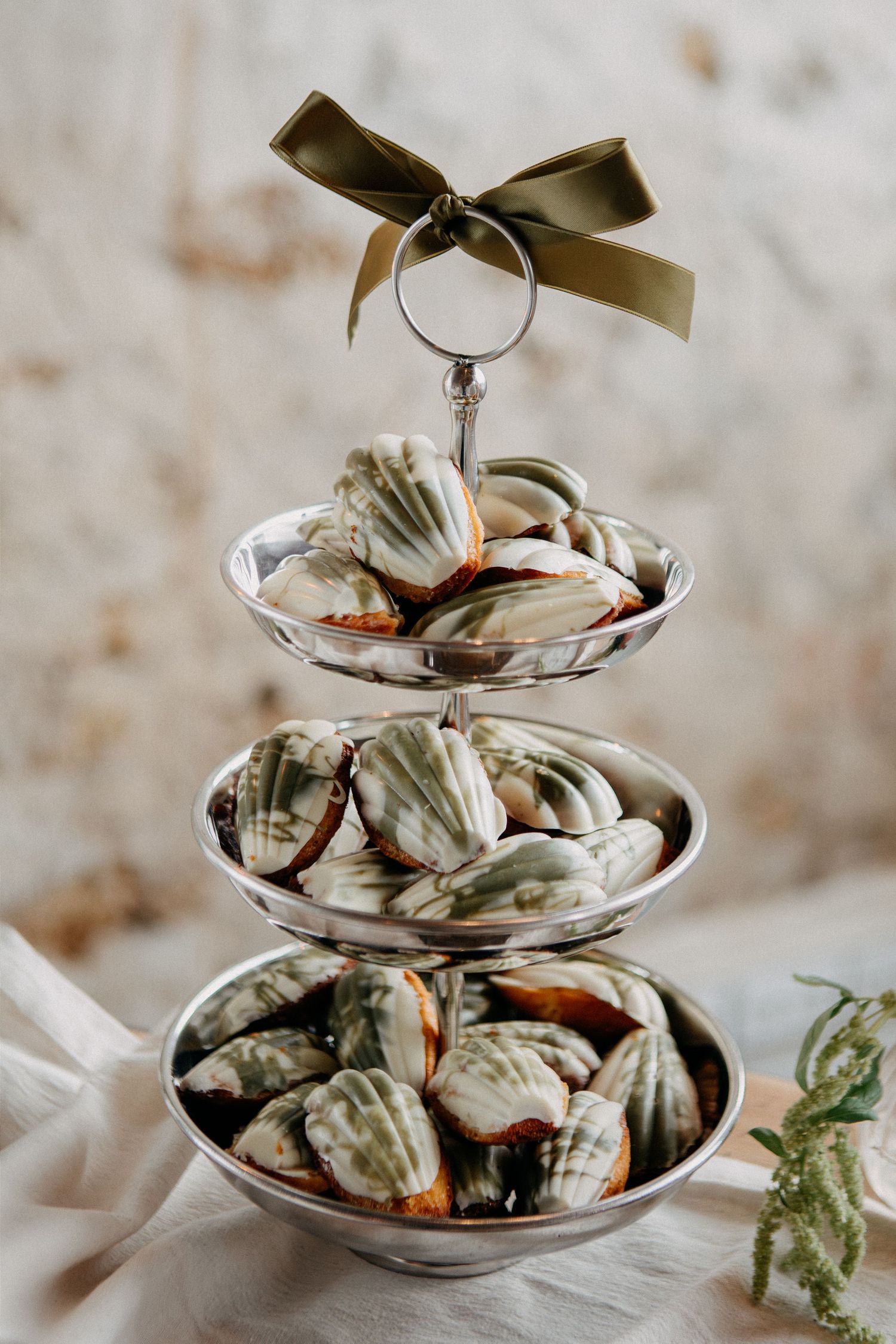 Three-tiered silver stand filled with green and brown marbled madeleines, topped with an olive green ribbon bow.