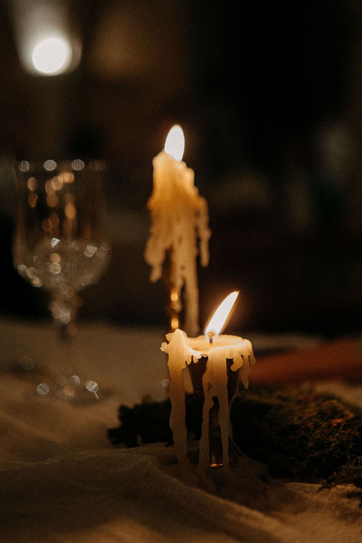 Two melting white candles glowing warmly on a dark table with a crystal glass in the background.