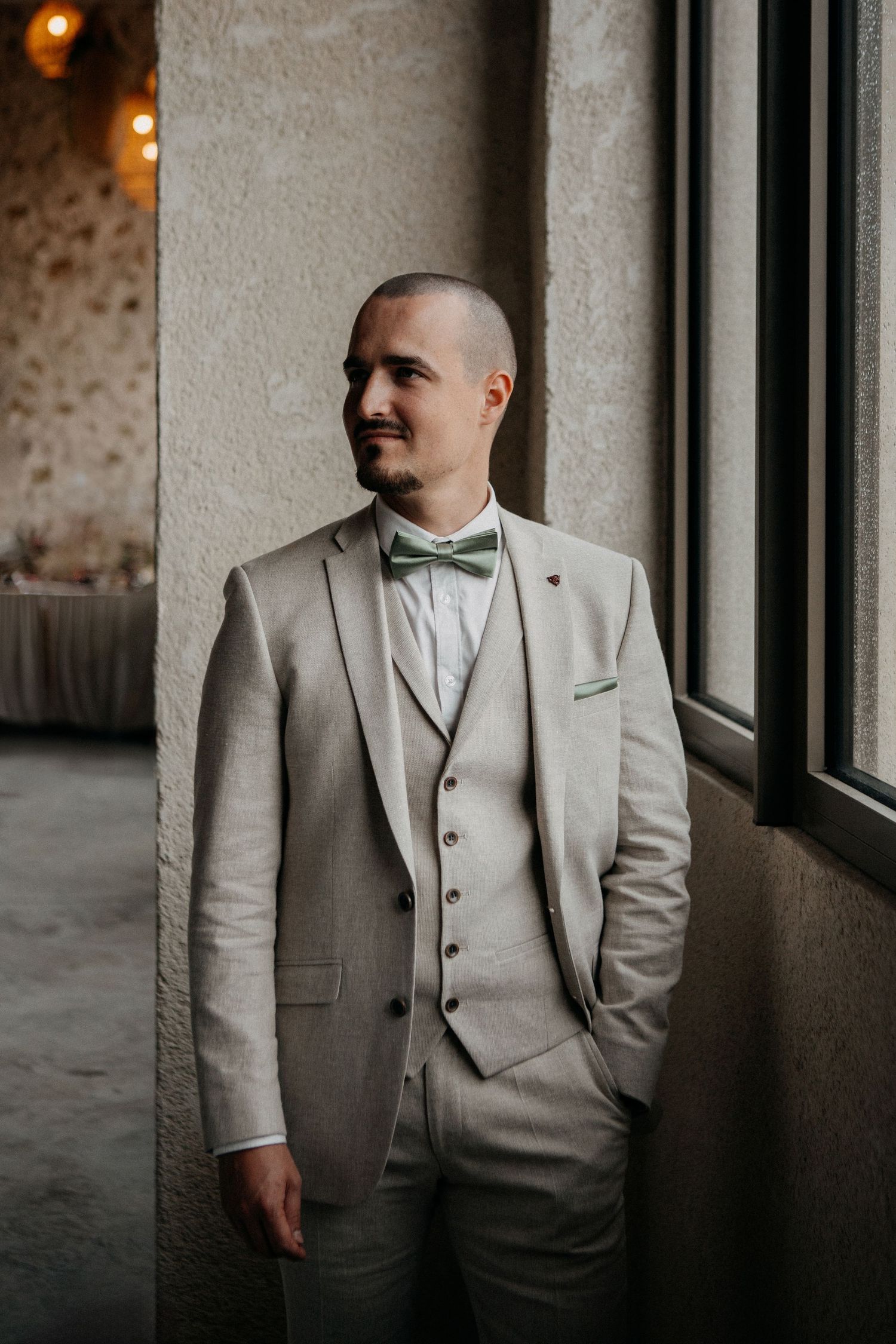 Dapper groom in beige three-piece suit with green bow tie posing by a window.