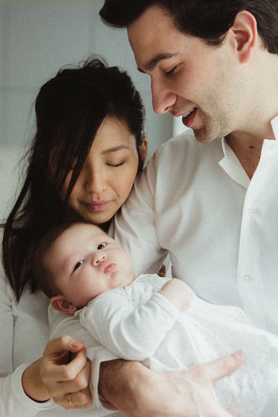 Happy parents in white shirts smiling at alert newborn baby they are holding.